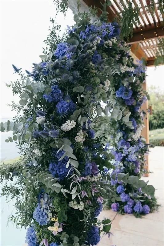 A floral arrangement with blue and white flowers and green foliage, arching over a wooden structure.