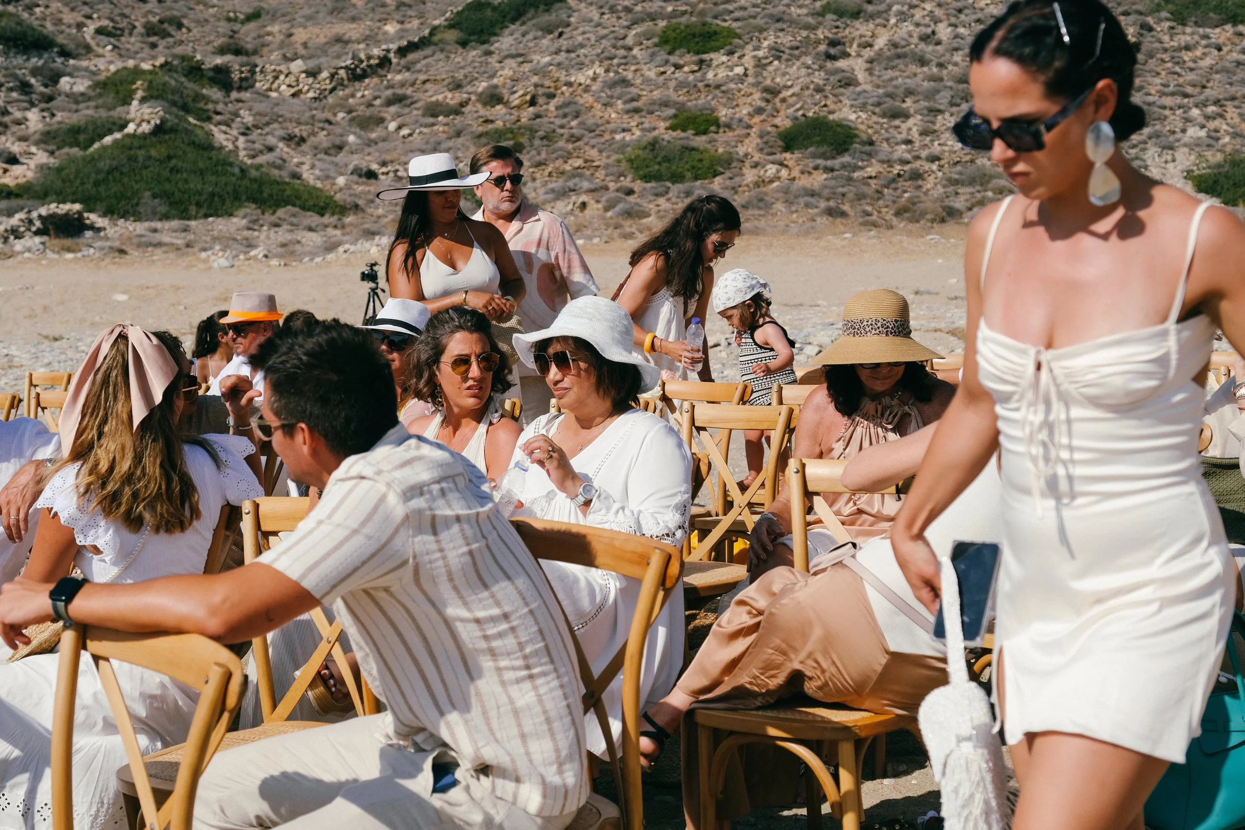 People sitting on wooden chairs outdoors on a beach with rocky hills in the background, many wearing summer dresses, hats, and sunglasses, suggesting a beach event or gathering.