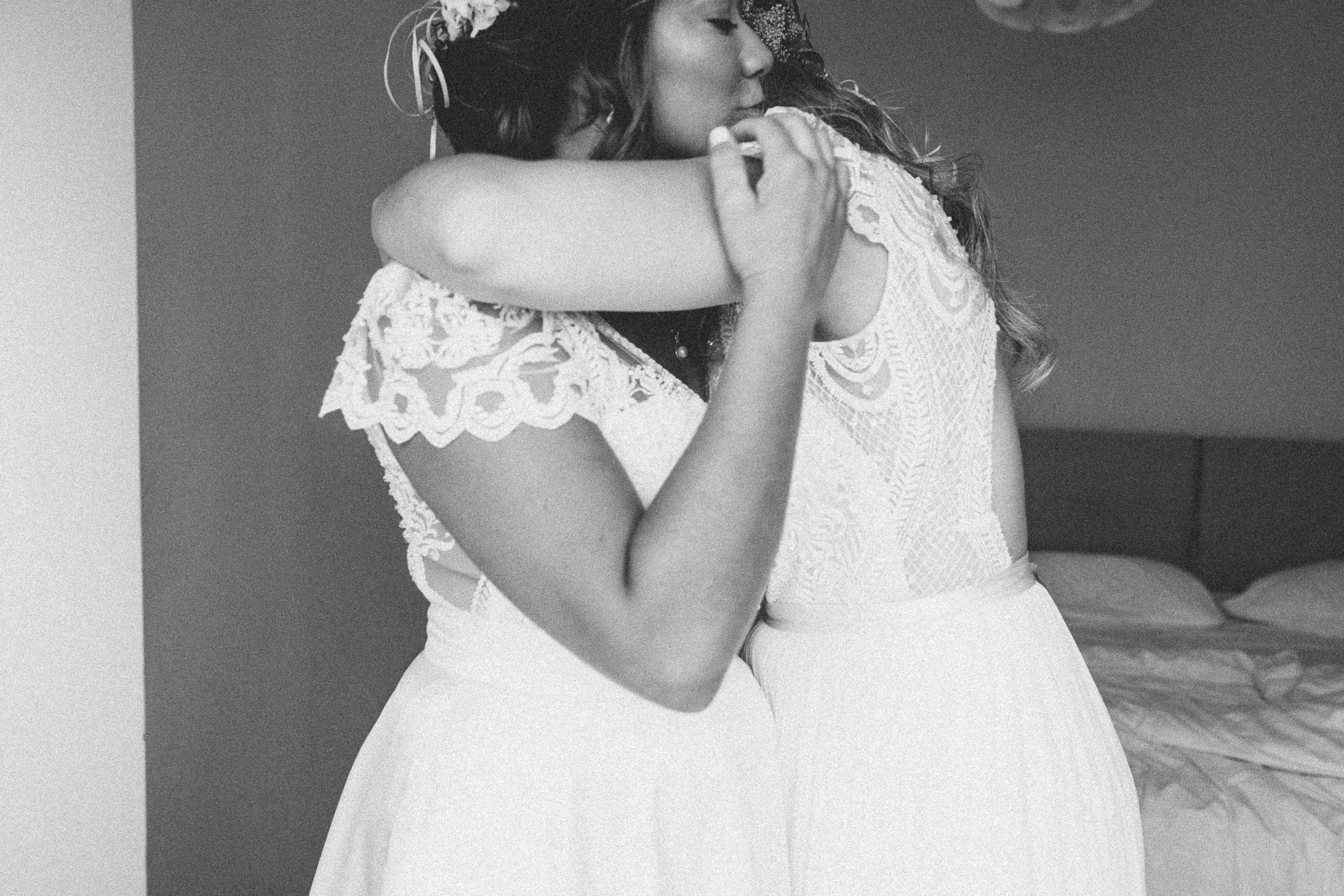 Two women dressed in white lace dresses hugging each other indoors, with a bed visible in the background.