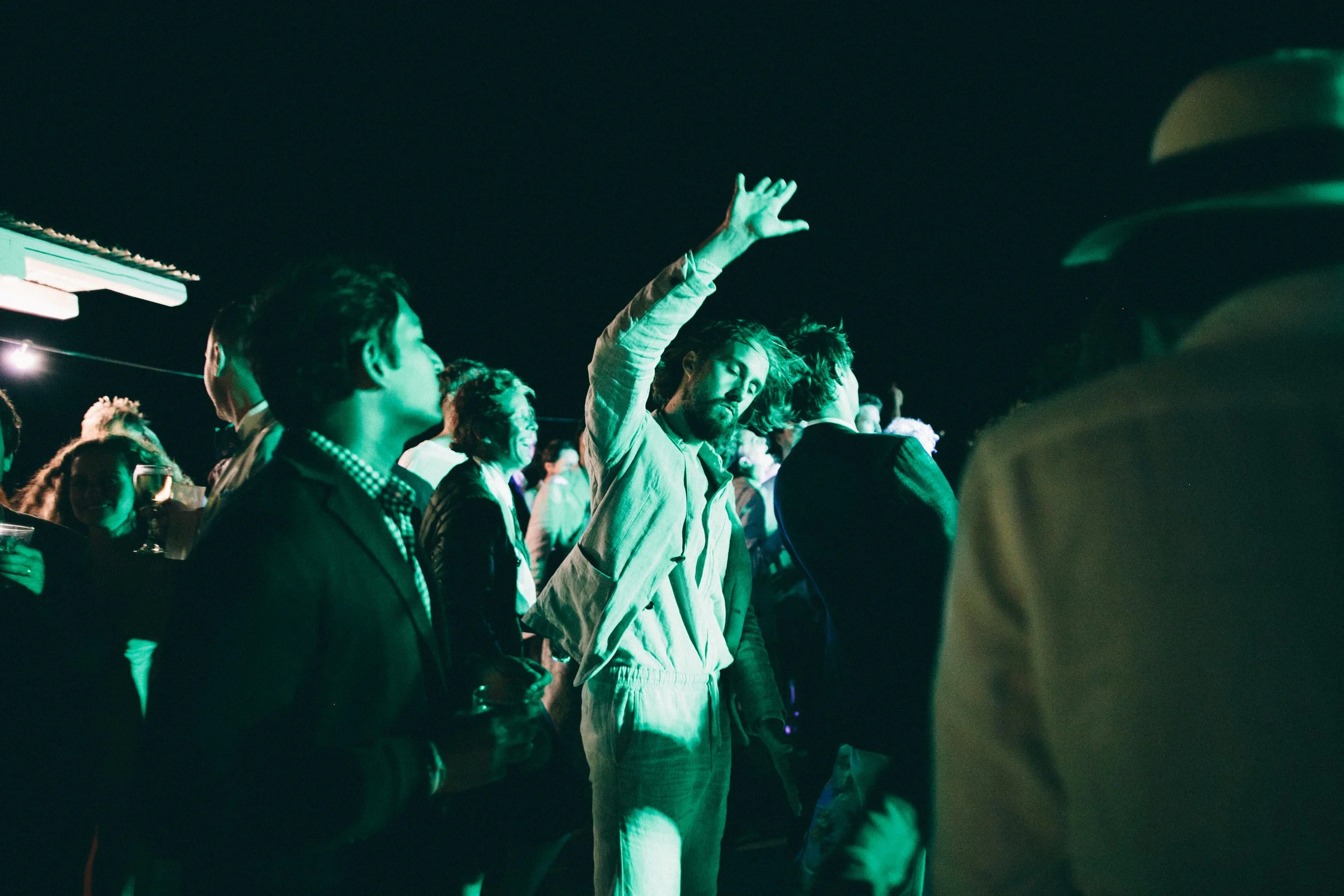 People attending an outdoor nightlife event, illuminated by green lighting, with one man raising his hand.