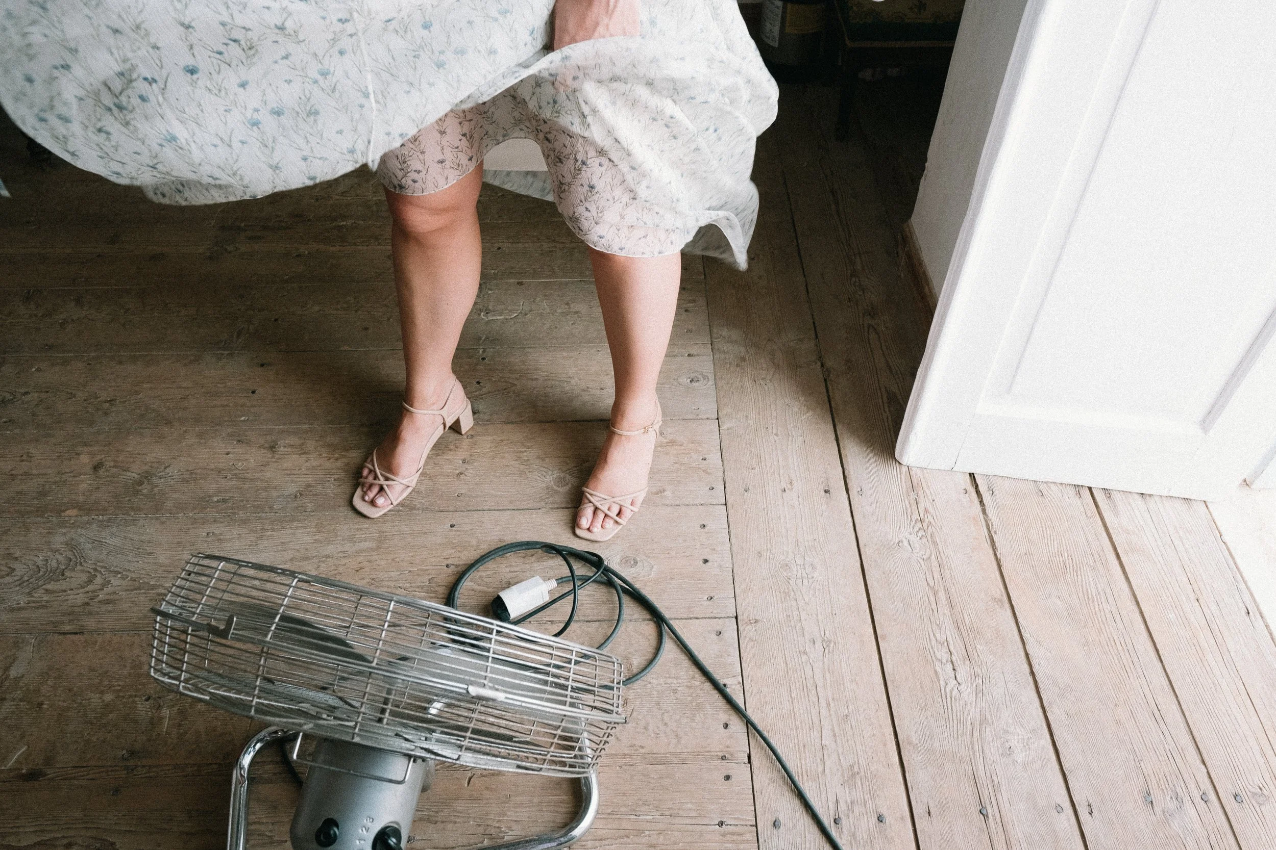 A woman wearing high heels and a patterned dress is standing in a room with wooden floorboards, next to a space heater and a door.