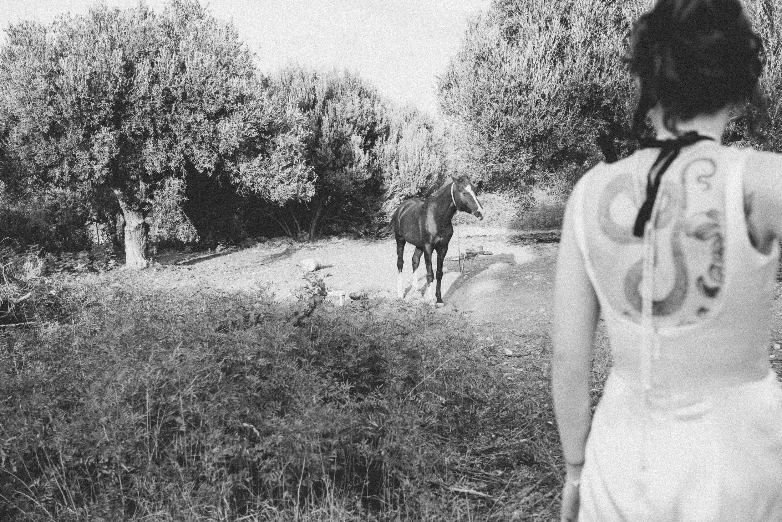 A woman with short hair wearing a tank top with a snake design on the back, standing outdoors, looking at a horse tied to a tree in a natural, bushy landscape.