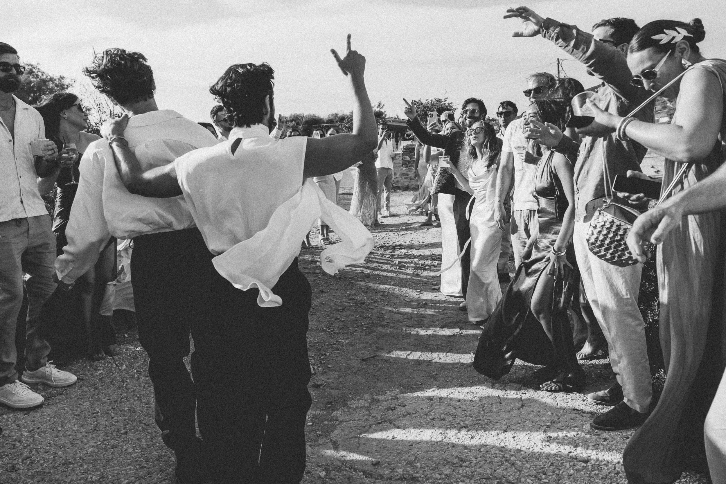 A black and white photo of a group of people at an outdoor gathering, some dancing and others standing, with a dirt ground and open sky.