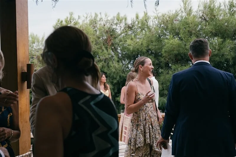 Group of people at an outdoor social gathering, with some conversing and a woman smiling while holding a drink, surrounded by greenery.