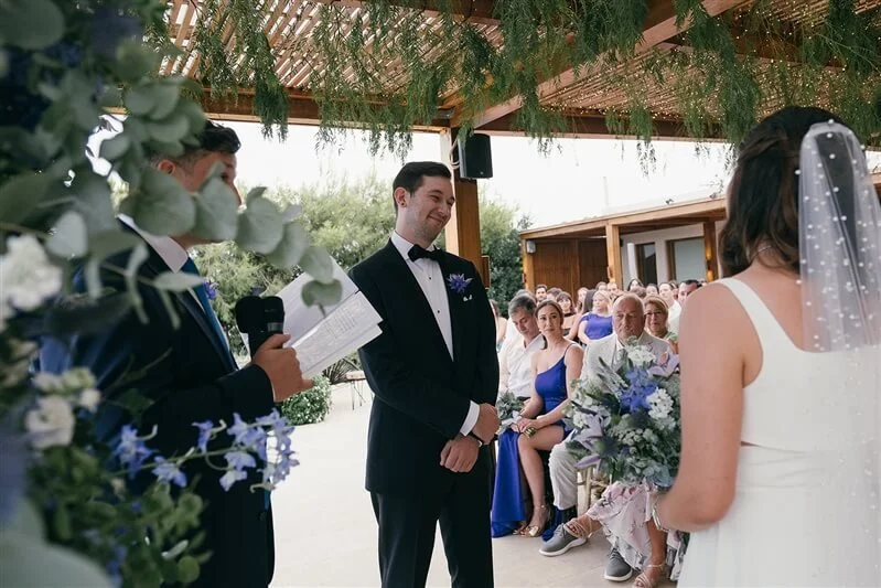A groom in a tuxedo with a bow tie looking at the bride in a white wedding dress and veil, during their outdoor wedding ceremony, with guests seated in the background.