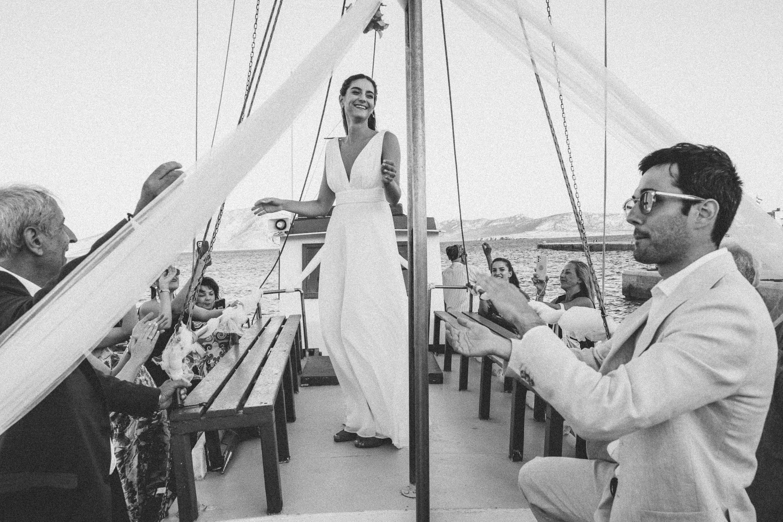 A woman in a white dress standing on a boat, smiling and talking to a man in a white suit who is sitting and using a tablet. Several people are taking pictures or watching, with water and mountains in the background.