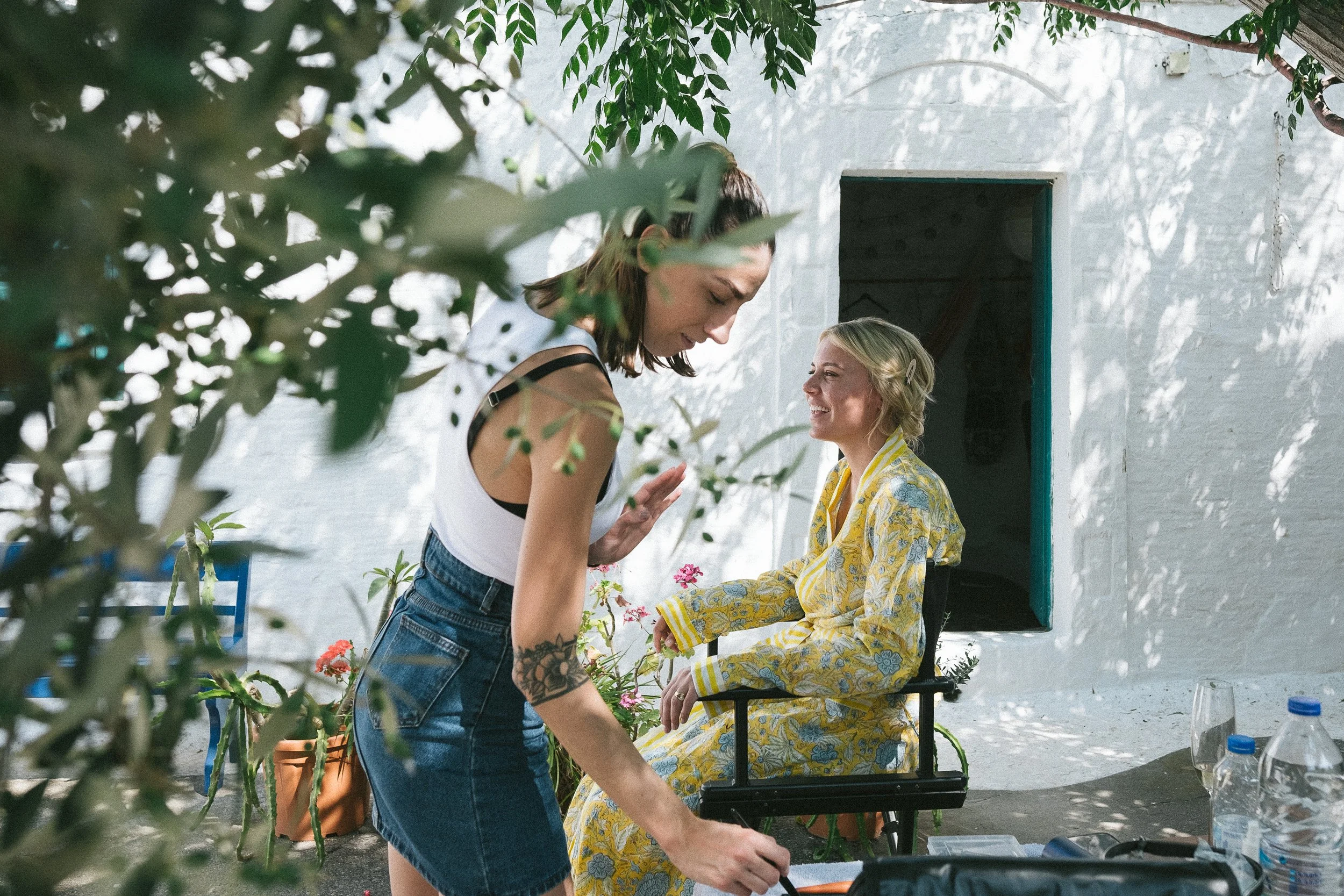 Two women engaging in conversation outdoors near a white wall with a window, surrounded by greenery, with bottles and glasses on a table.