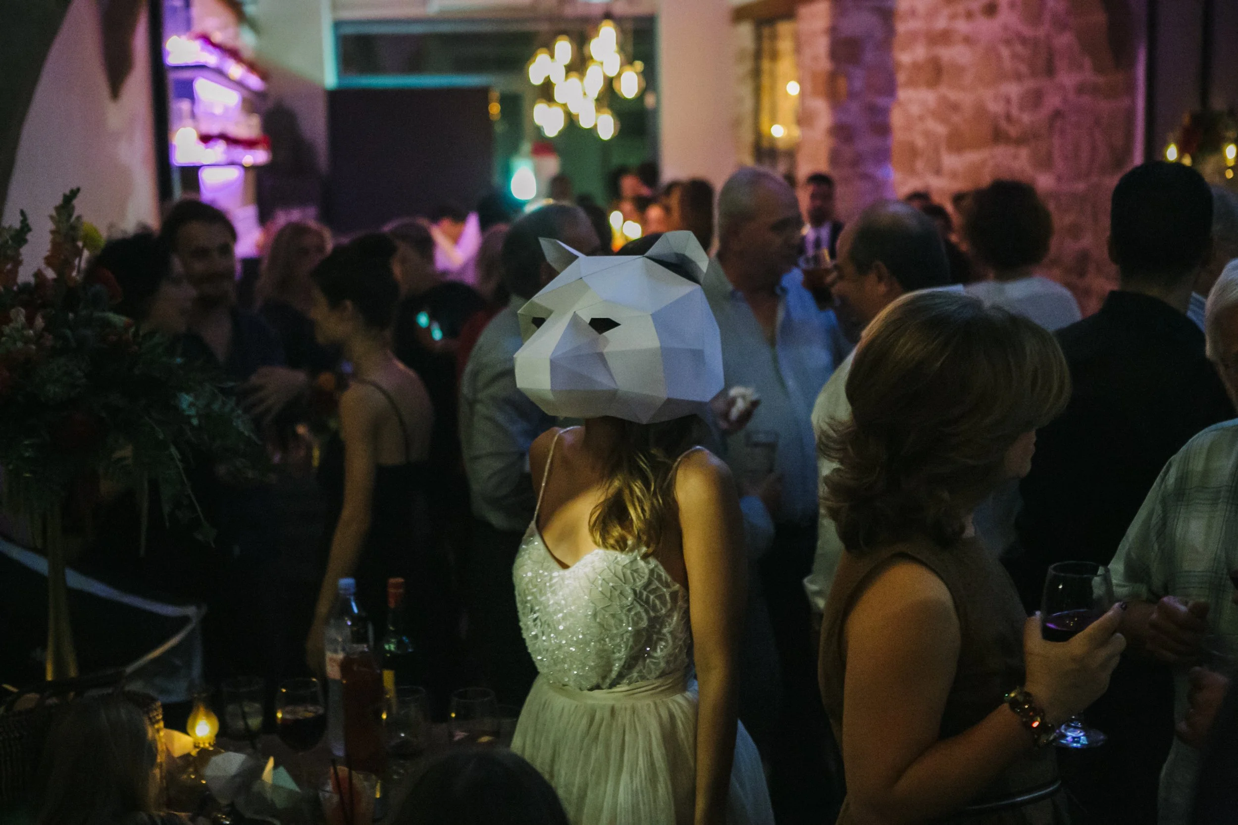 A woman at a party wearing a large, white, polygonal animal mask, possibly a fox or wolf. She is dressed in a sleeveless, light-colored dress with thin straps, and the scene is dimly lit with other guests socializing and drinking in the background.