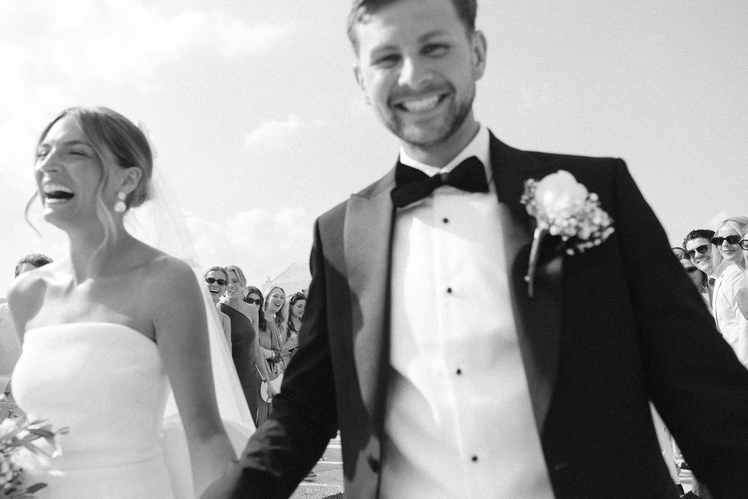 Black-and-white photo of a wedding celebration. The bride and groom are smiling and holding hands, surrounded by a group of guests who are also smiling and wearing sunglasses.