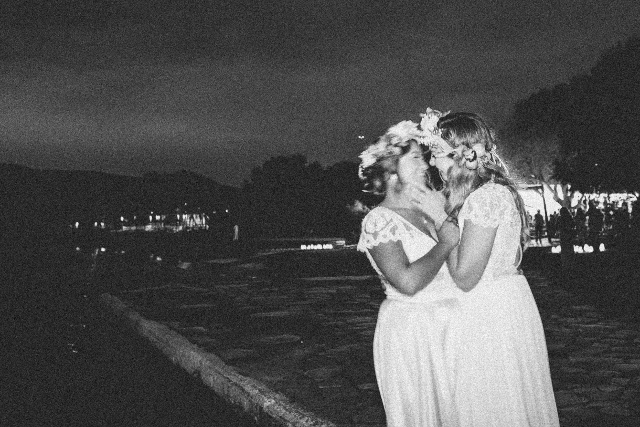 A black and white photo of two women in lace dresses embracing each other on a waterfront at night, with trees, a dock, and people in the background.
