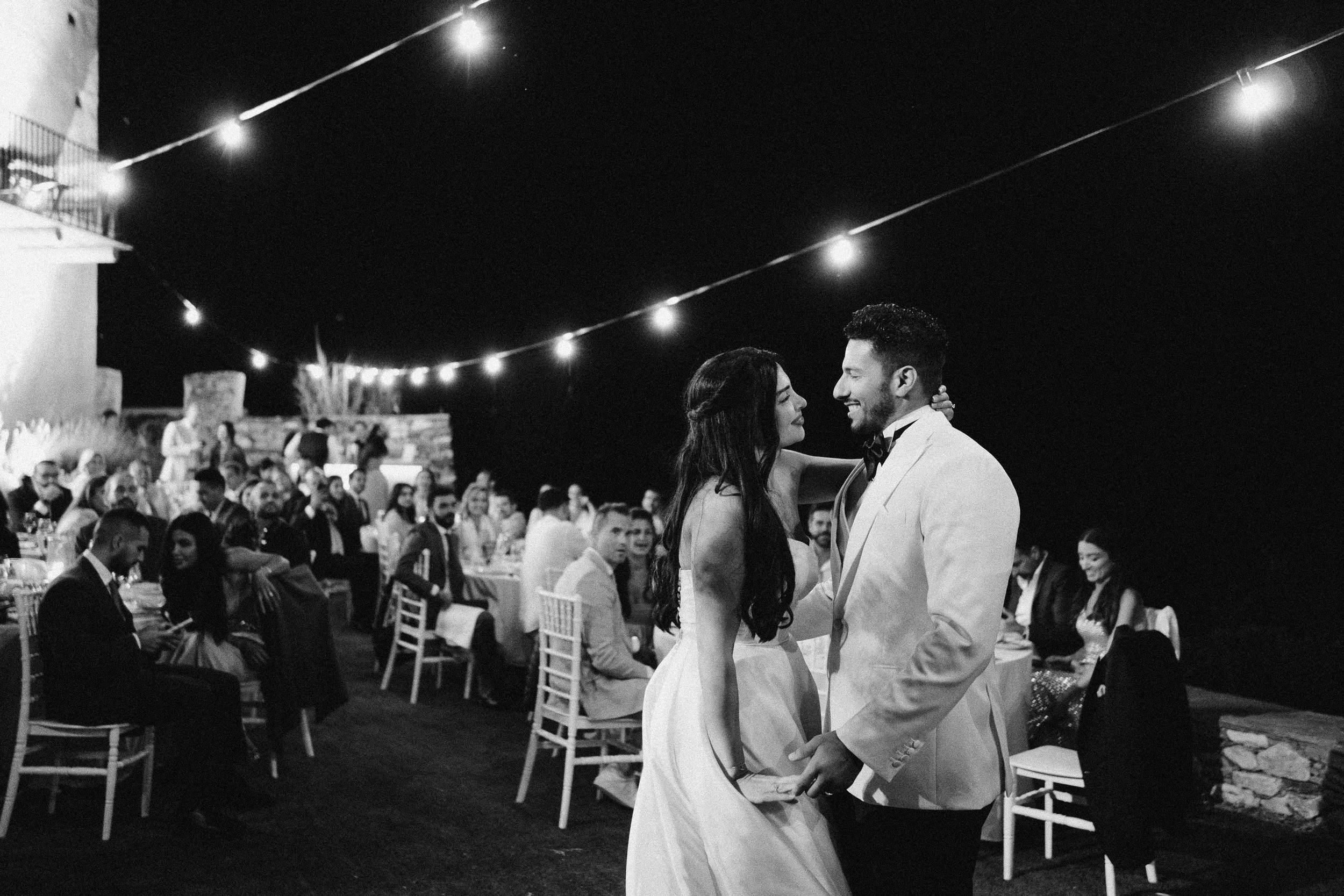 A black and white photo of a wedding reception with the bride and groom dancing together, surrounded by seated guests at outdoor tables under string lights at night.