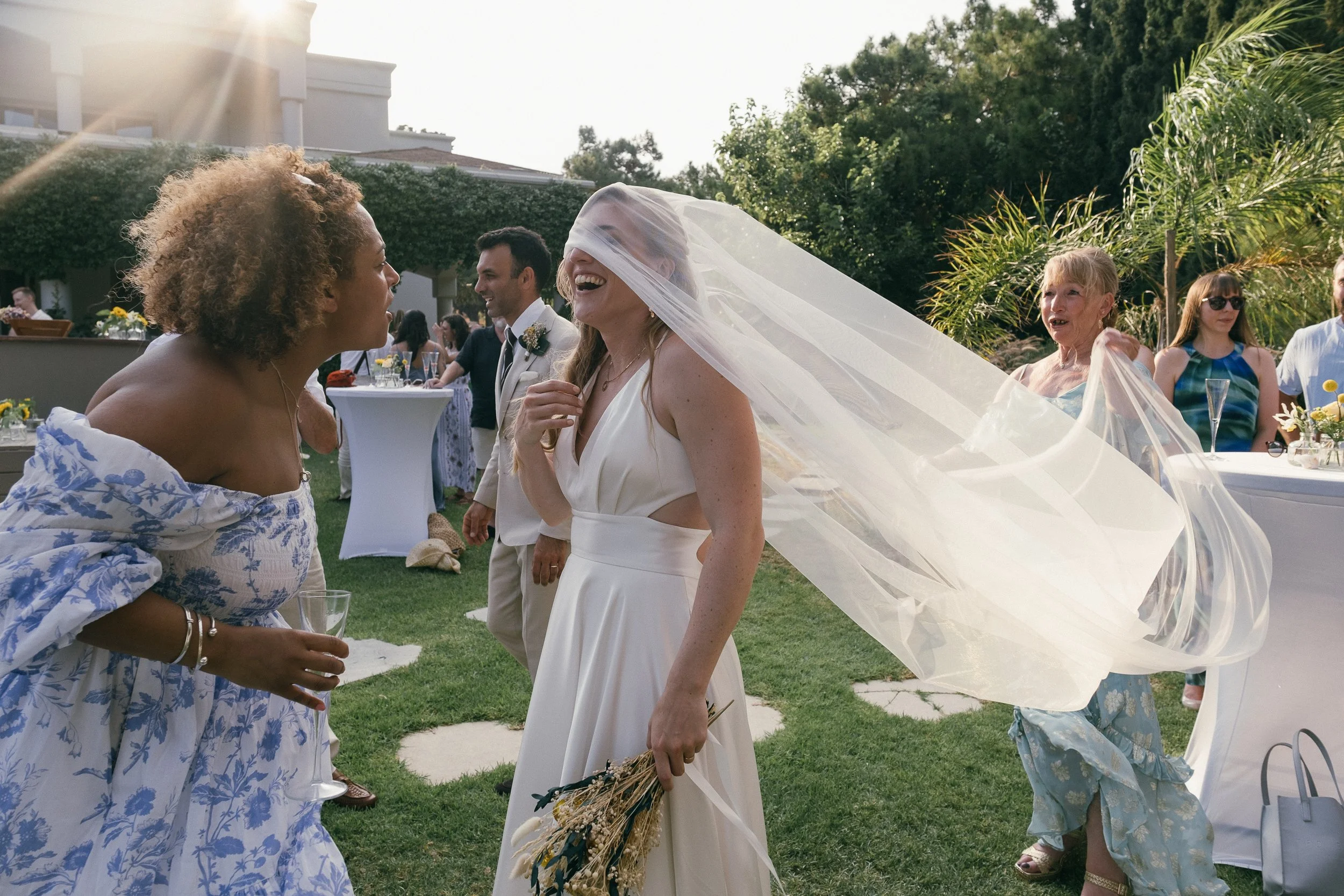 A bride in a white dress and veil laughing and chatting with a woman in a blue and white floral dress at an outdoor wedding reception, with guests and greenery in the background.