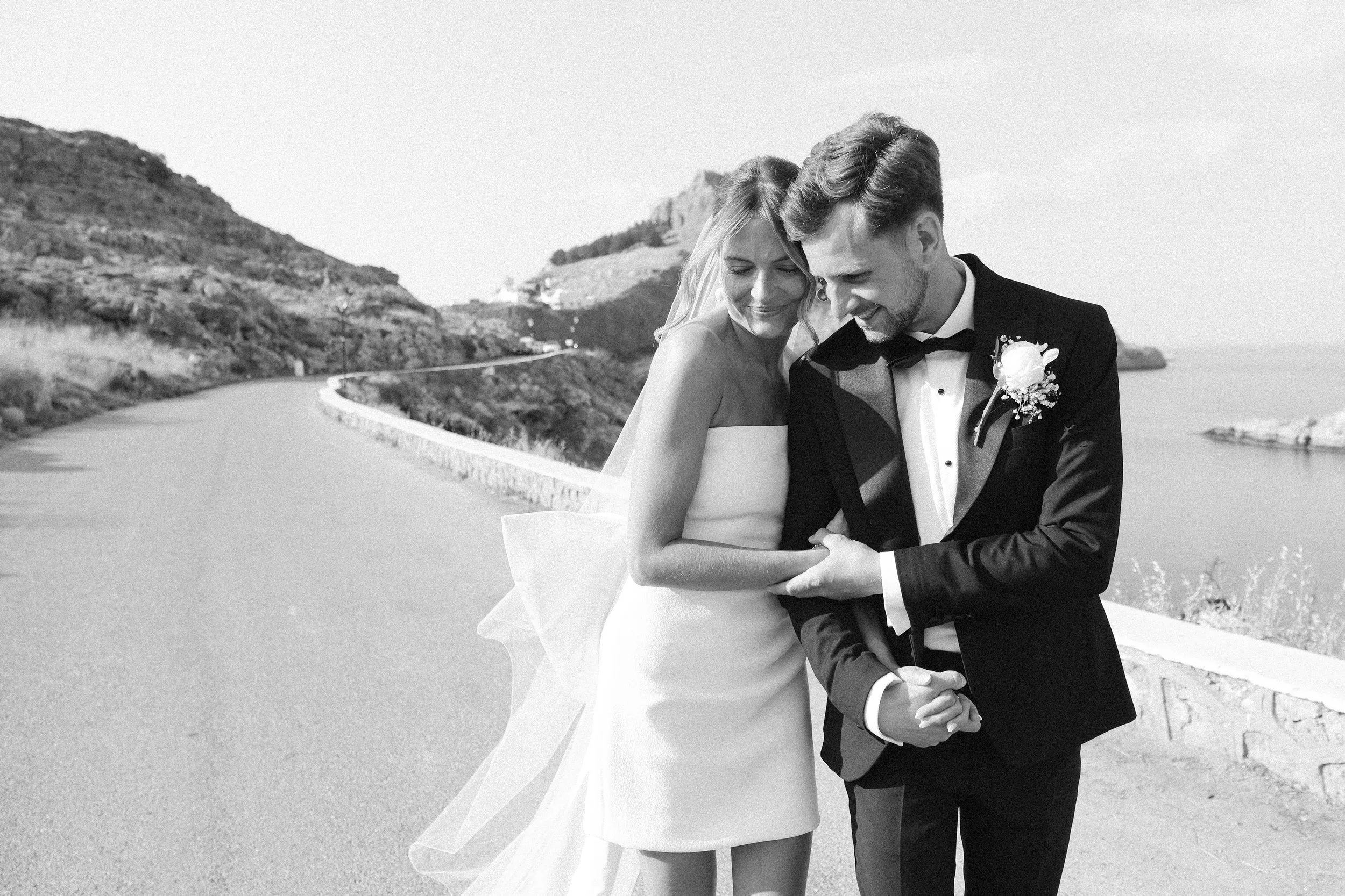 Black and white photograph of a newlywed couple walking along a scenic coastal road. The bride is wearing a strapless dress with a veil, and the groom is dressed in a tuxedo with a boutonniere. They are smiling and holding hands, with hills and the o