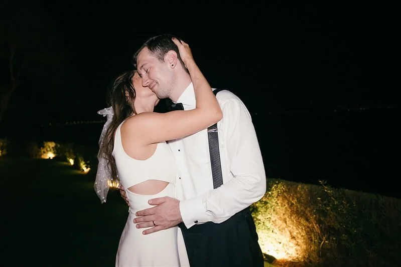 A couple in formal attire kissing outdoors at night, illuminated by soft lighting.