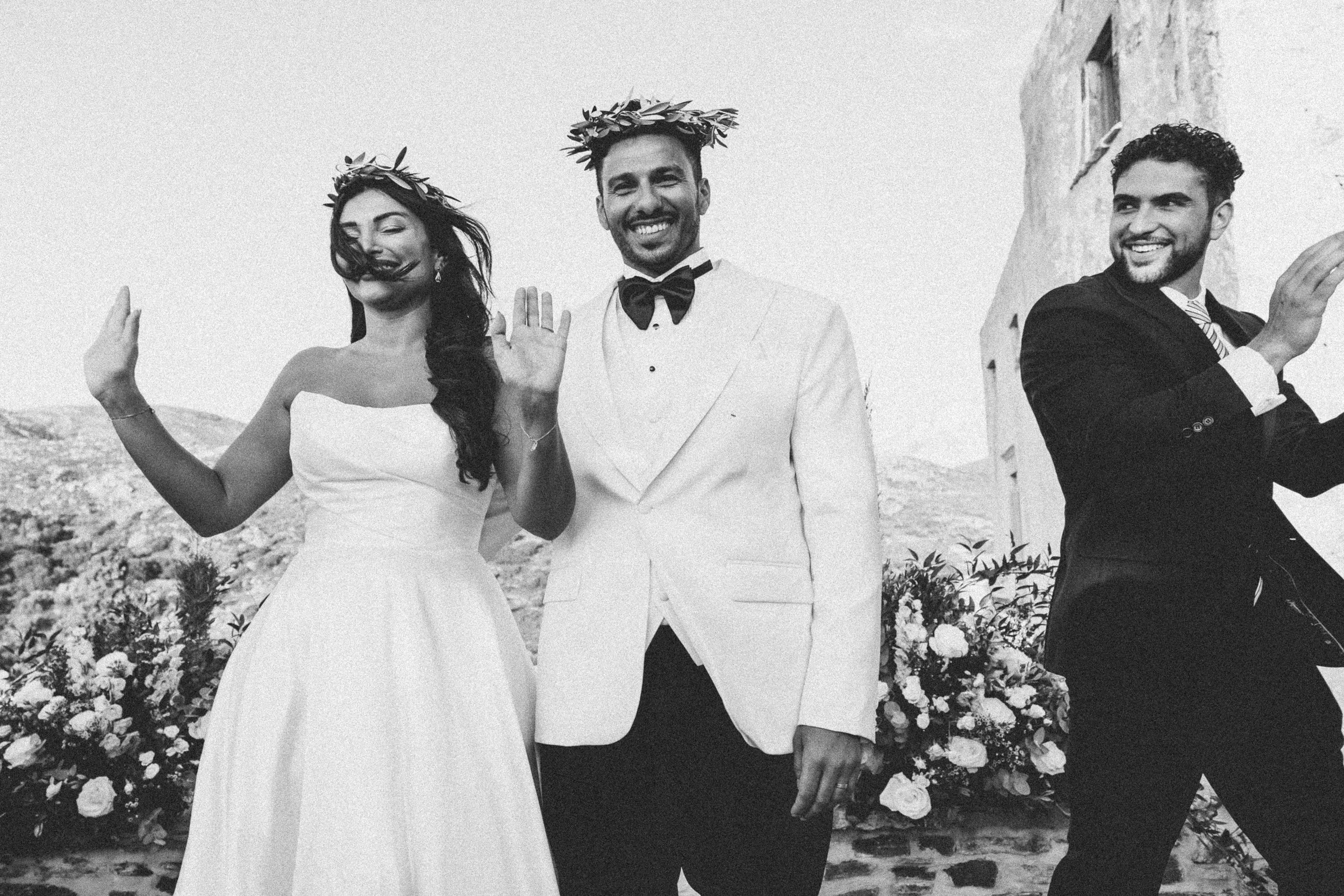 Black and white photo of a wedding ceremony with a bride, groom, and a man celebrating outdoors with mountains and flowers in the background.