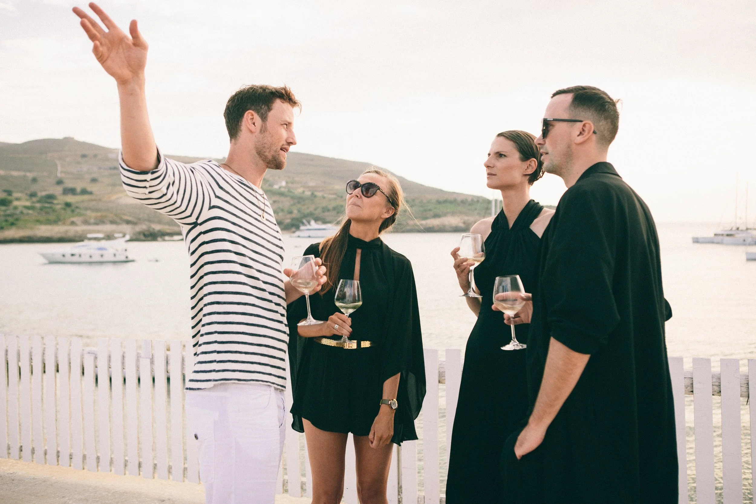 Four people having a conversation by the waterfront, holding wine glasses, with boats and hills in the background.