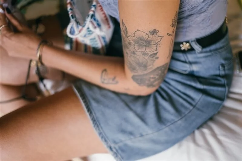 Close-up of a person's arm with tattoos, wearing a gray t-shirt and denim shorts, sitting on a bed with a colorful bag nearby.