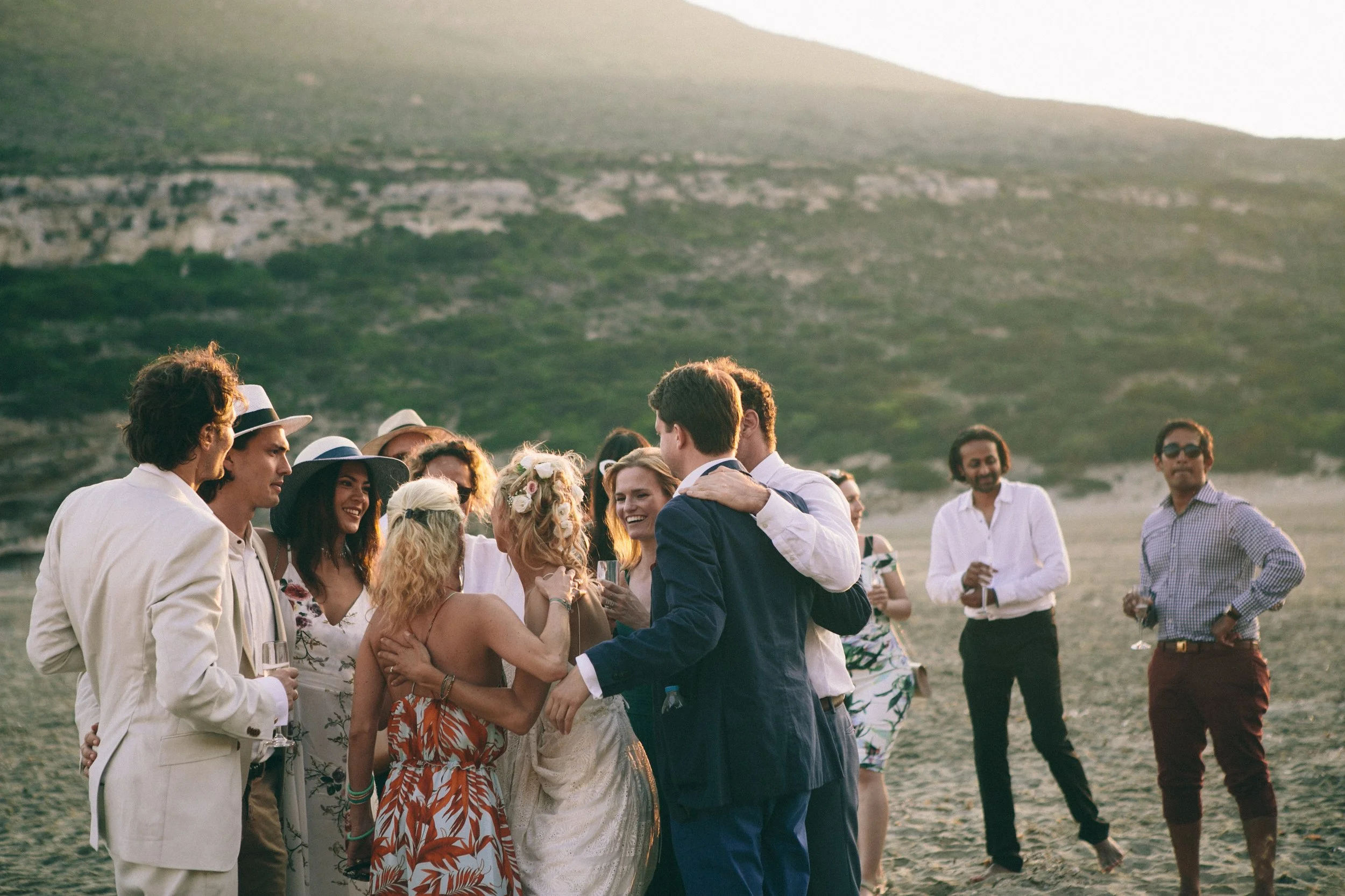 A group of people dressed in formal and semi-formal attire, gathered on a beach at sunset, celebrating and socializing.