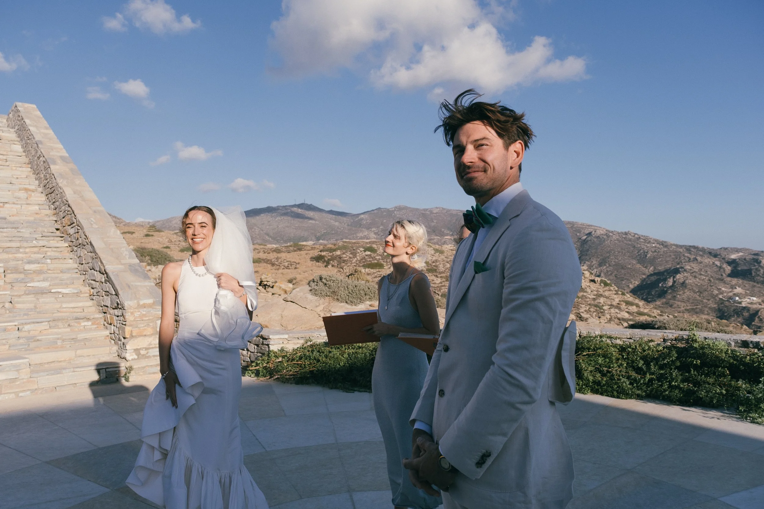A wedding ceremony outdoors with three people, a bride in a white dress and veil, a groom in a light suit, and a female officiant holding a book, set against a mountainous landscape with a blue sky.