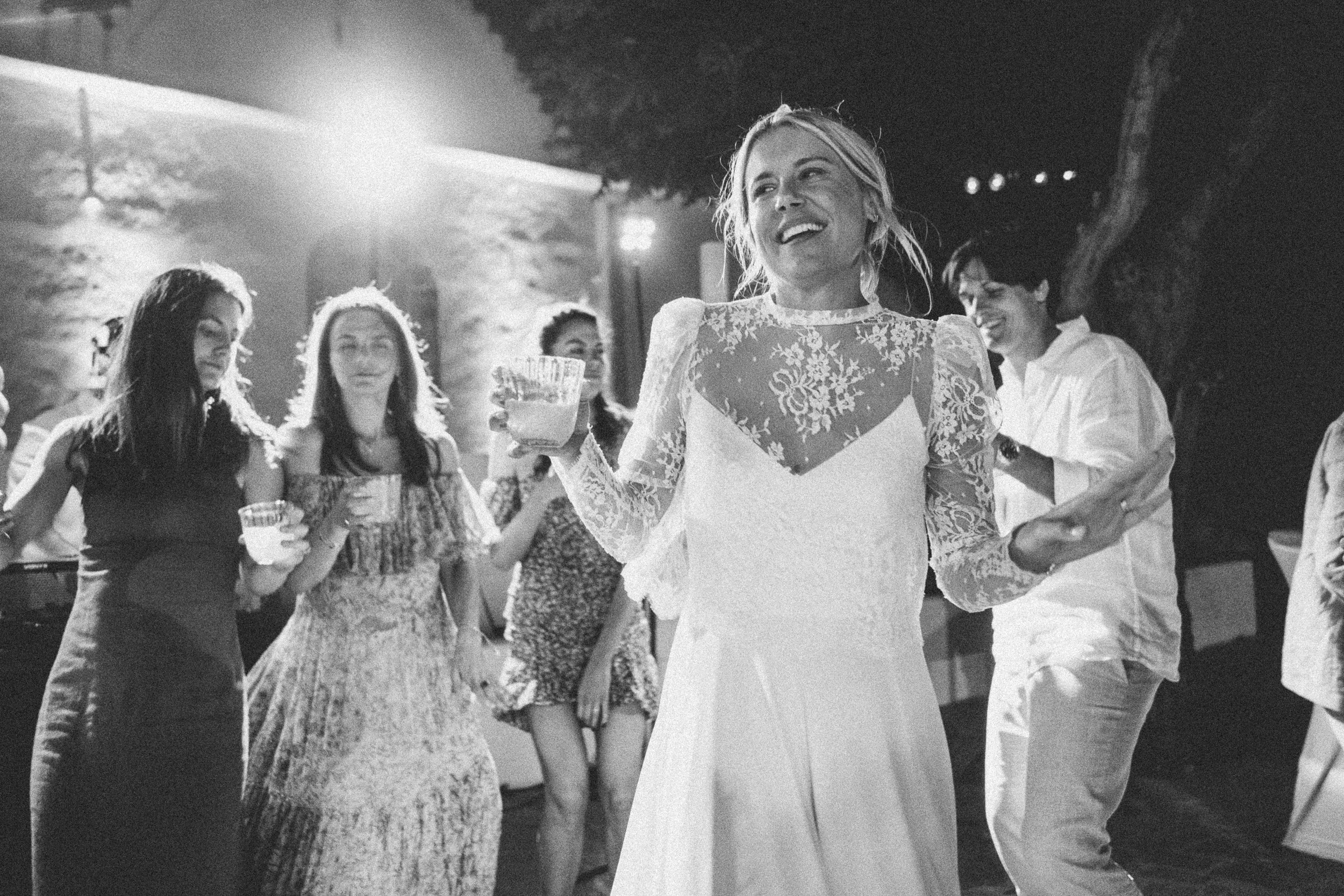 Black and white photo of a woman in a wedding dress holding a drink, dancing and smiling, surrounded by people at a party or celebration outdoors at night.
