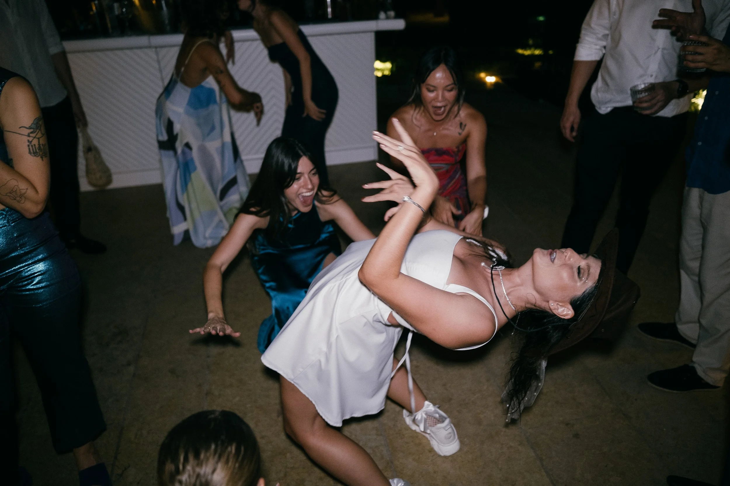 Group of women dancing and having fun at a party, one woman in a white dress and hat is dancing in the center, while others are surrounding her and clapping.