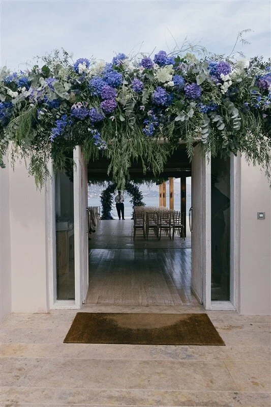 Wedding ceremony space with a floral arch above the entrance, view of chairs, and a person standing near a body of water outside.