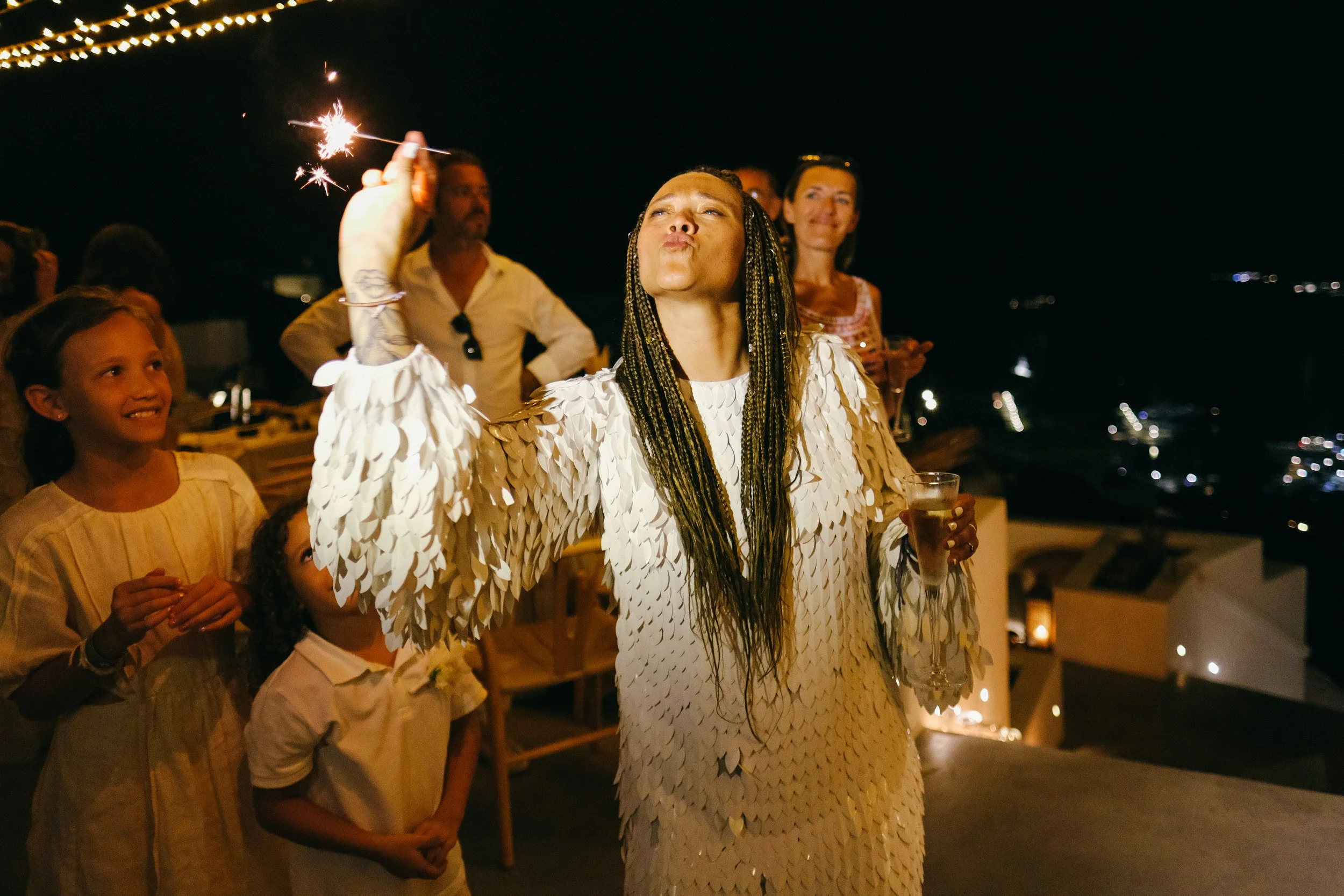 Woman with long braids wearing a white feathered dress holding a sparkler and a glass at night, surrounded by smiling people at a celebration with string lights and dark sky in the background.