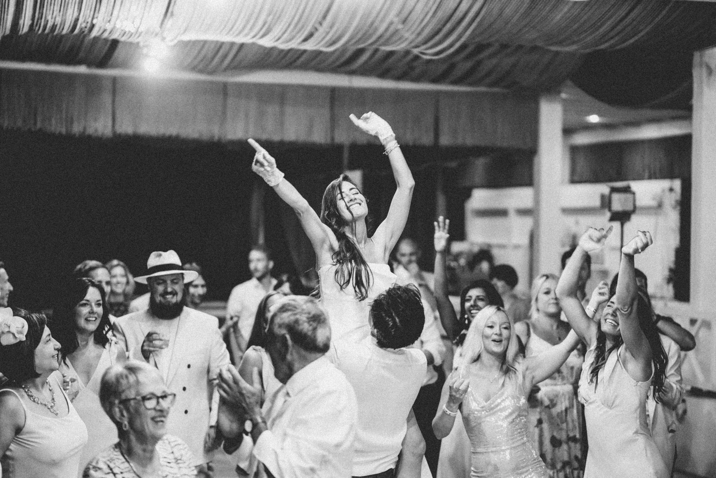 A woman in a wedding dress is being lifted by a man, surrounded by smiling guests at a celebration or wedding reception, lively dancing and joy evident.
