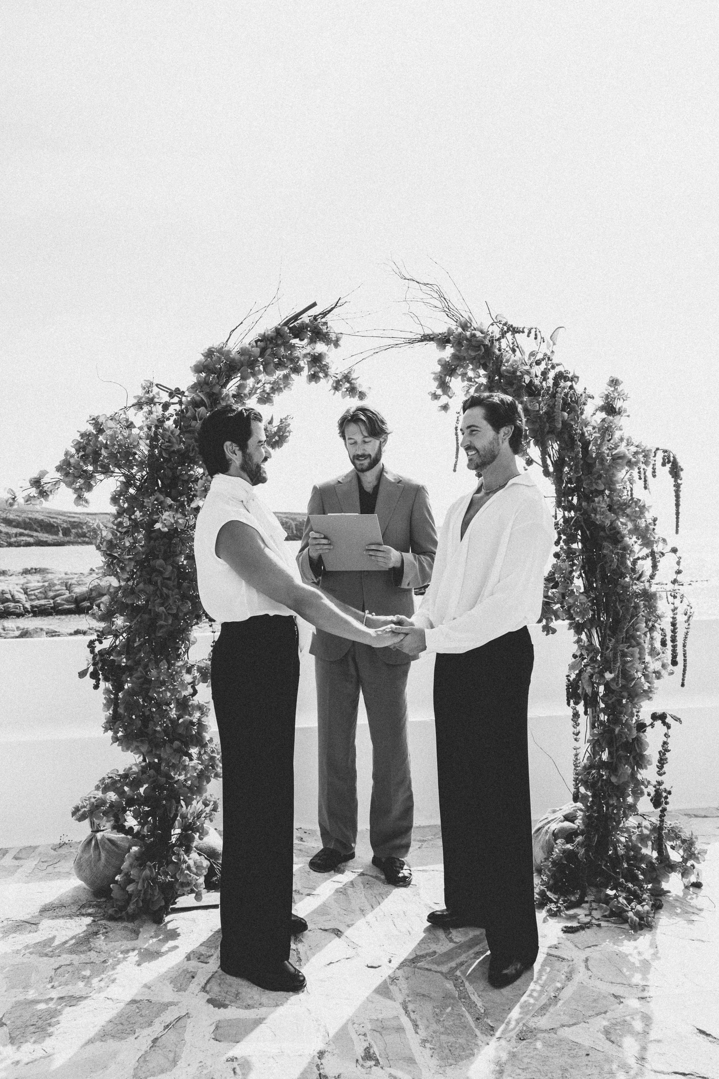 Two men holding hands during a wedding ceremony in front of a floral arch on a sunny day, with an officiant standing behind them, outdoors near the water.