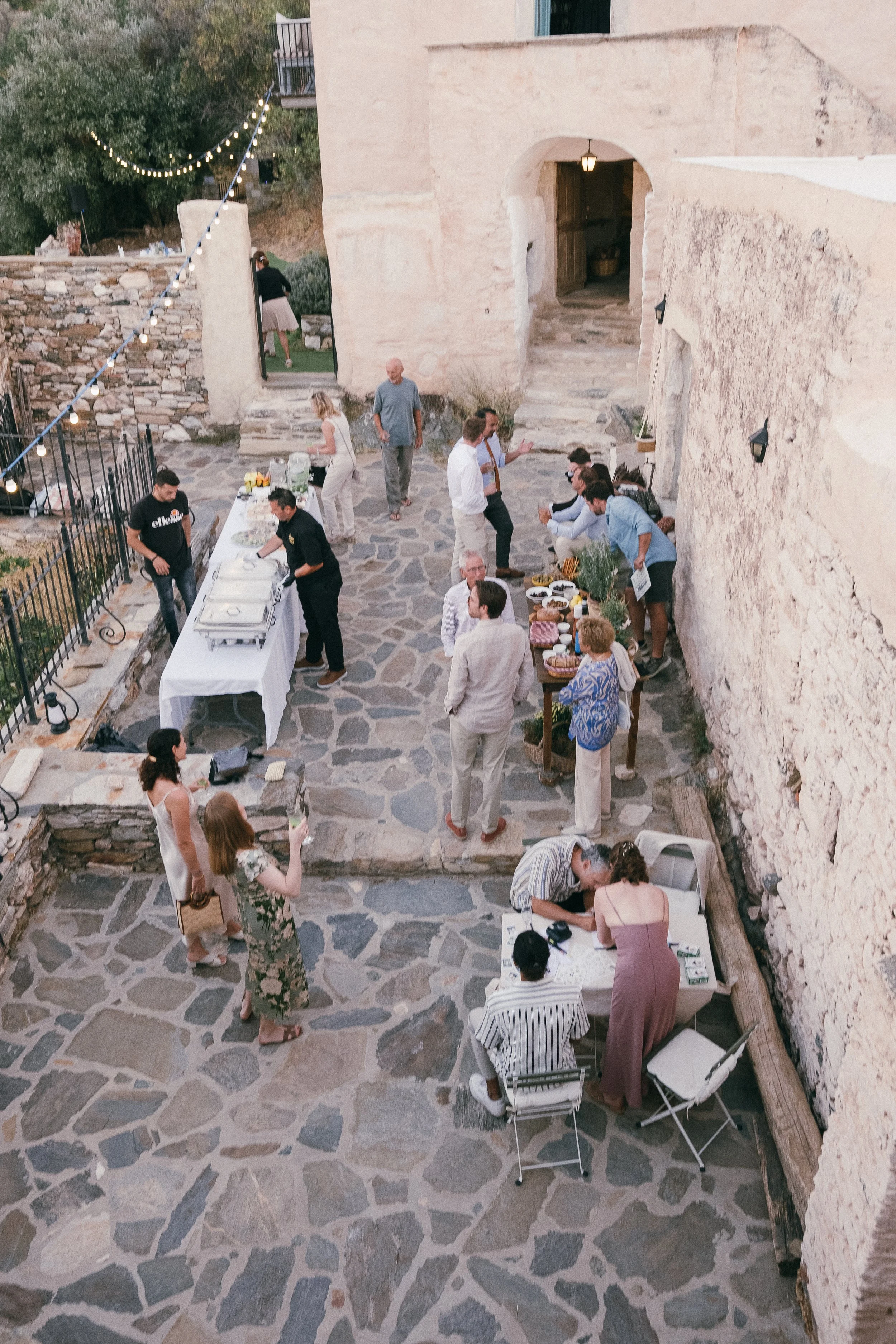 People gathering at an outdoor evening event with food tables, string lights, and a stone courtyard with rustic walls.
