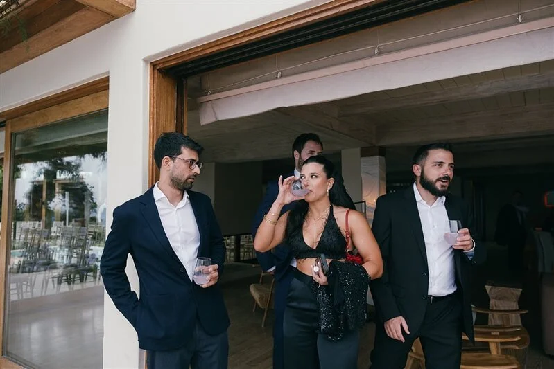 Group of four professionally dressed people at a social gathering, two men in suits and a woman in a black dress drinking and chatting indoors.