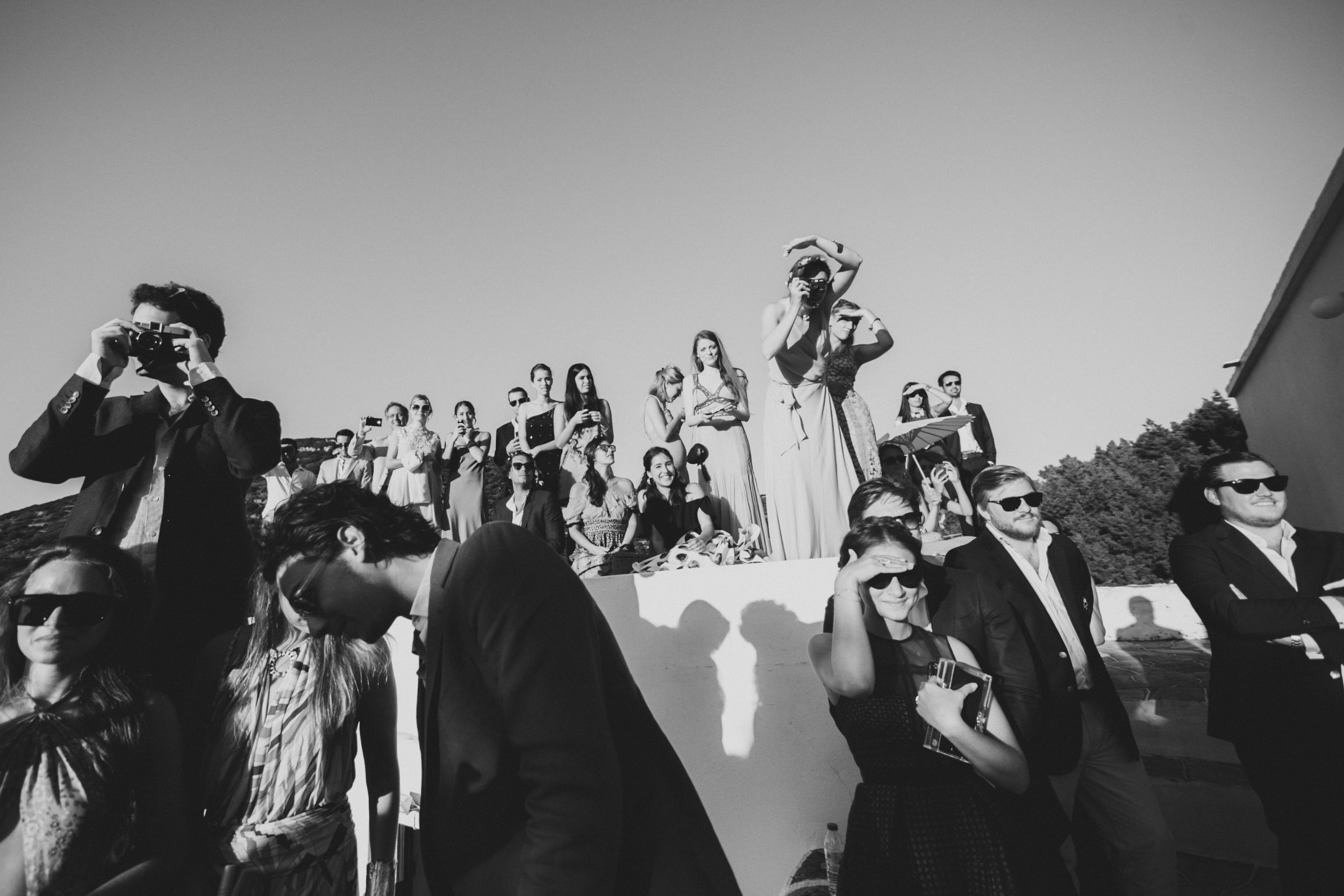 Black and white photo of a crowd of people, many wearing sunglasses, gathered outdoors on a sunny day, some taking photos or looking in different directions.