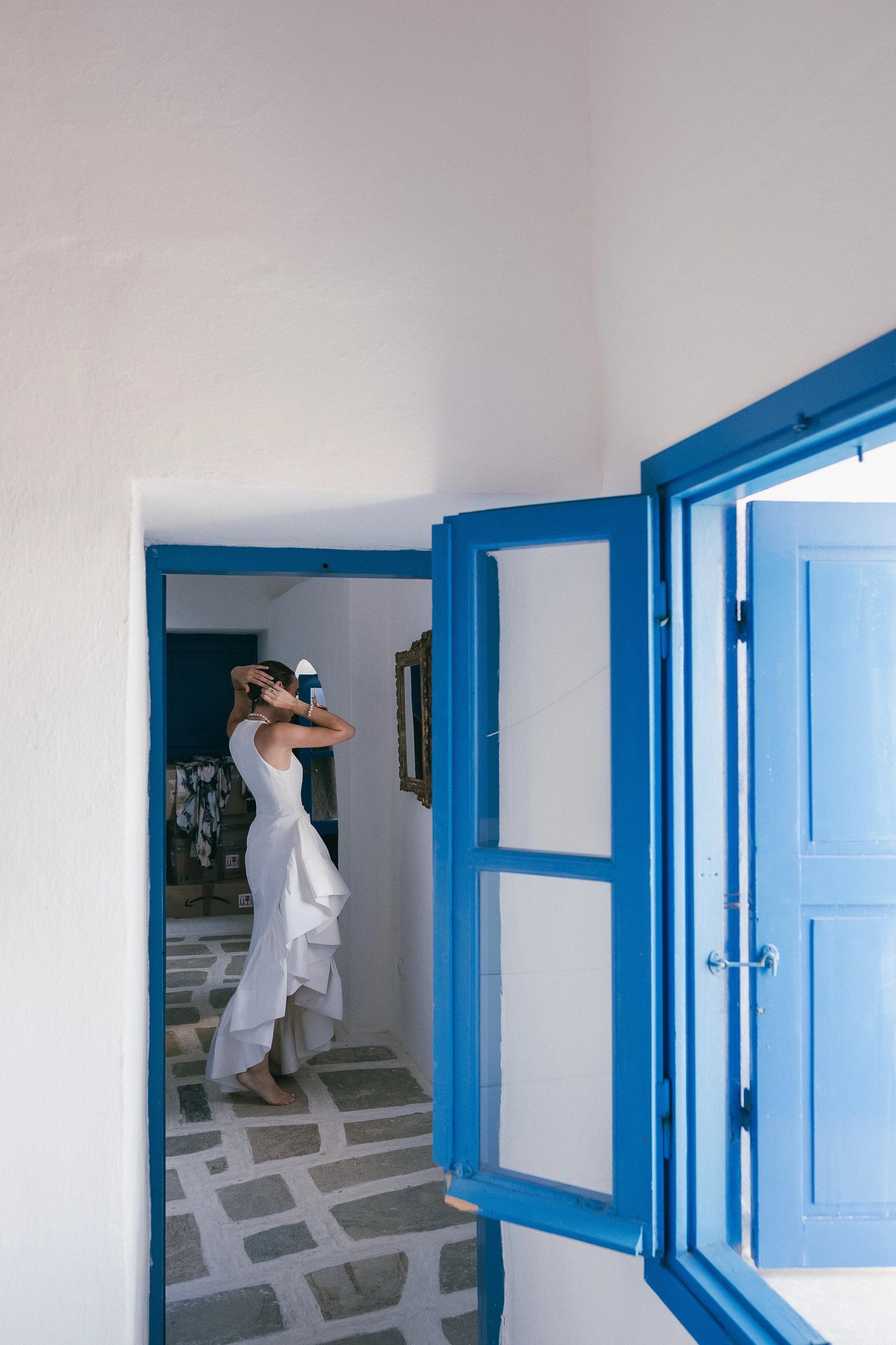 A woman in a white dress is standing in front of a mirror, adjusting her hair, as seen through an open blue window in a white-walled room.