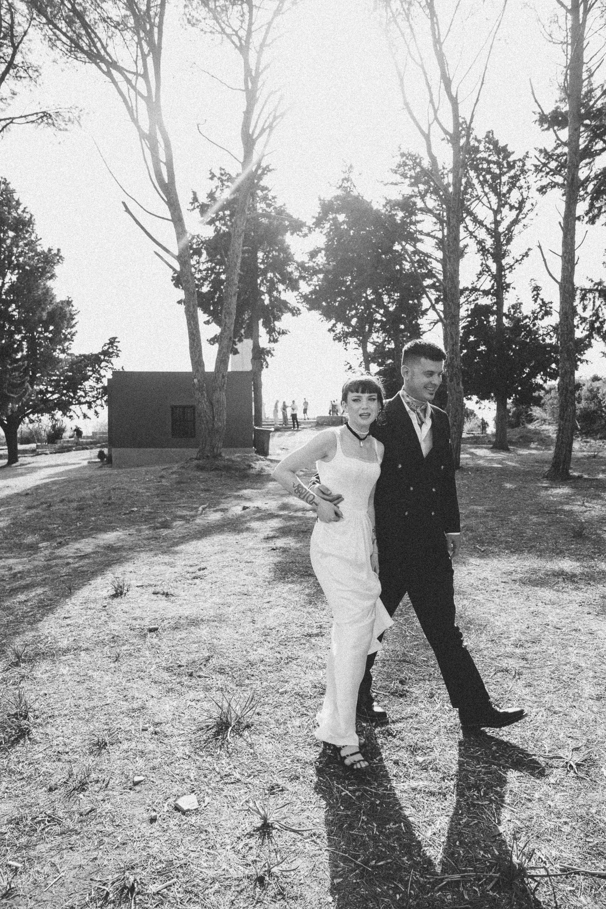 A black-and-white photo of a couple walking outdoors, with the woman wearing a white dress and the man in a dark suit. They are walking on a dirt path surrounded by trees, with sunlight filtering through the branches.