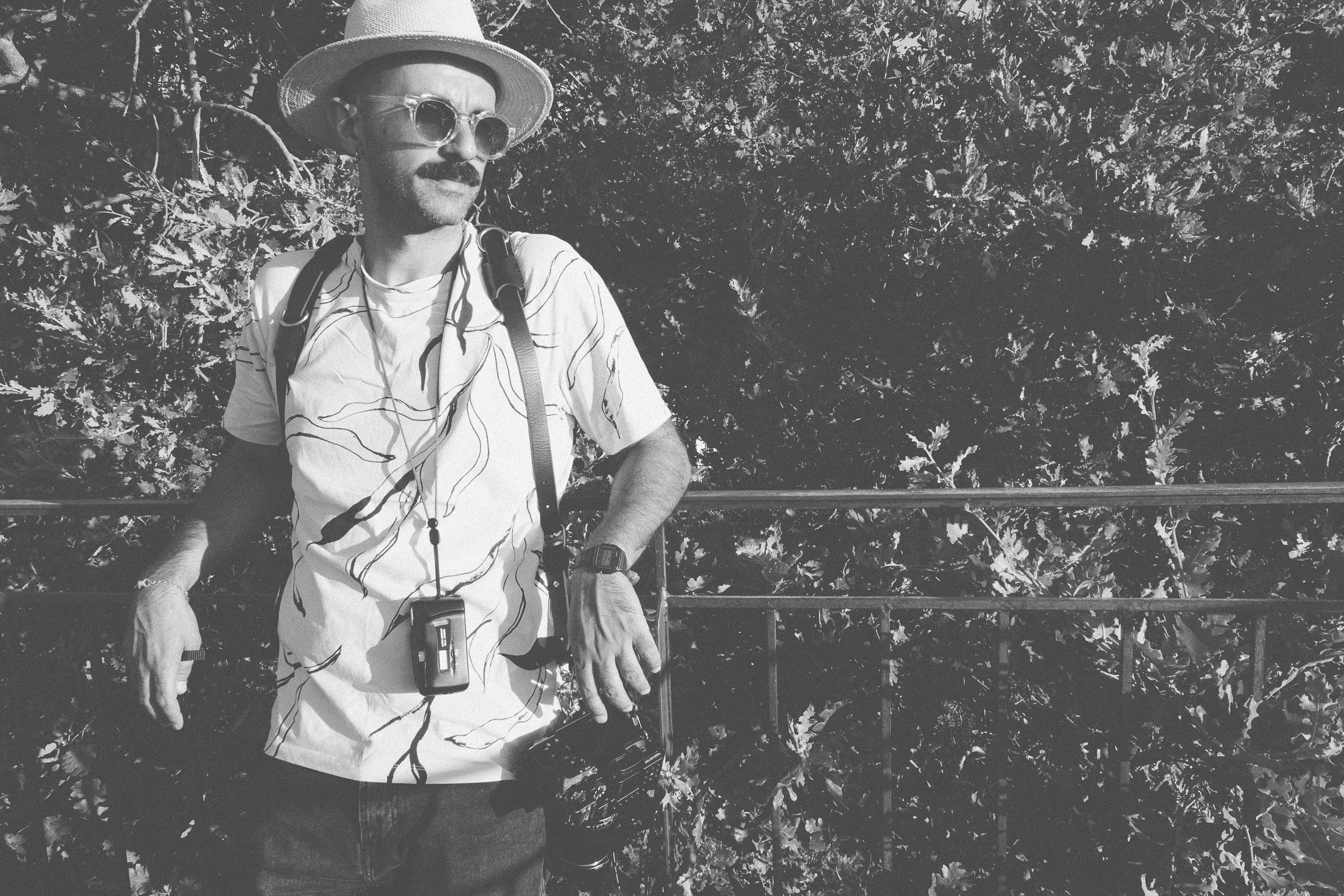 A man with a mustache wearing sunglasses, a wide-brimmed hat, a patterned T-shirt, and a backpack standing outdoors against a leafy background, holding a camera.