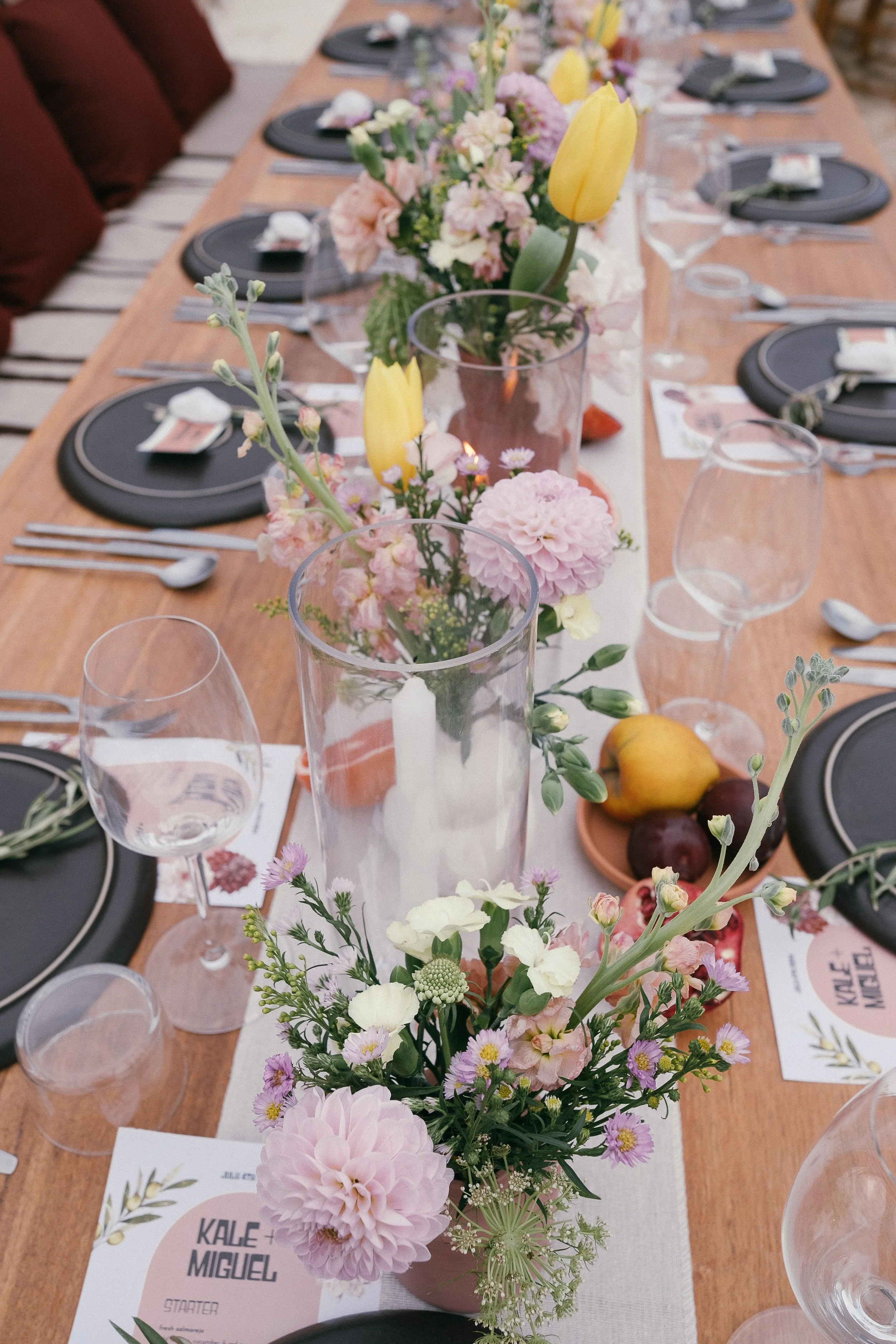 A decorated dining table with black plates, clear wine glasses, floral centerpieces with pink, yellow, and white flowers, candles, and small plates of fruit including pears and plums, set for a gathering.