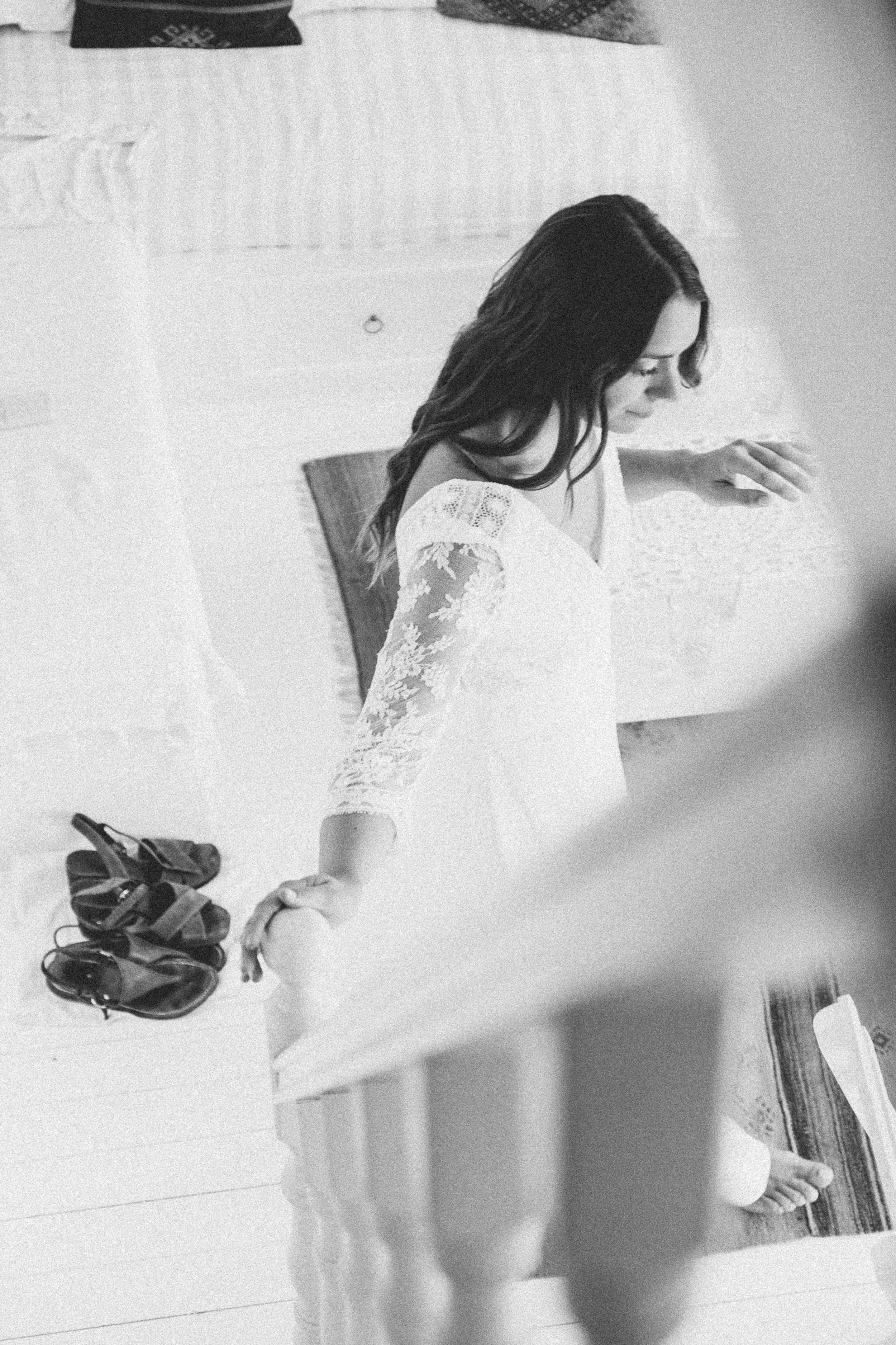 Black and white photo of a woman in a lace dress, possibly a wedding dress, standing on a staircase, looking down. Shoes are placed on the floor to the left, with a handrail visible in the foreground.