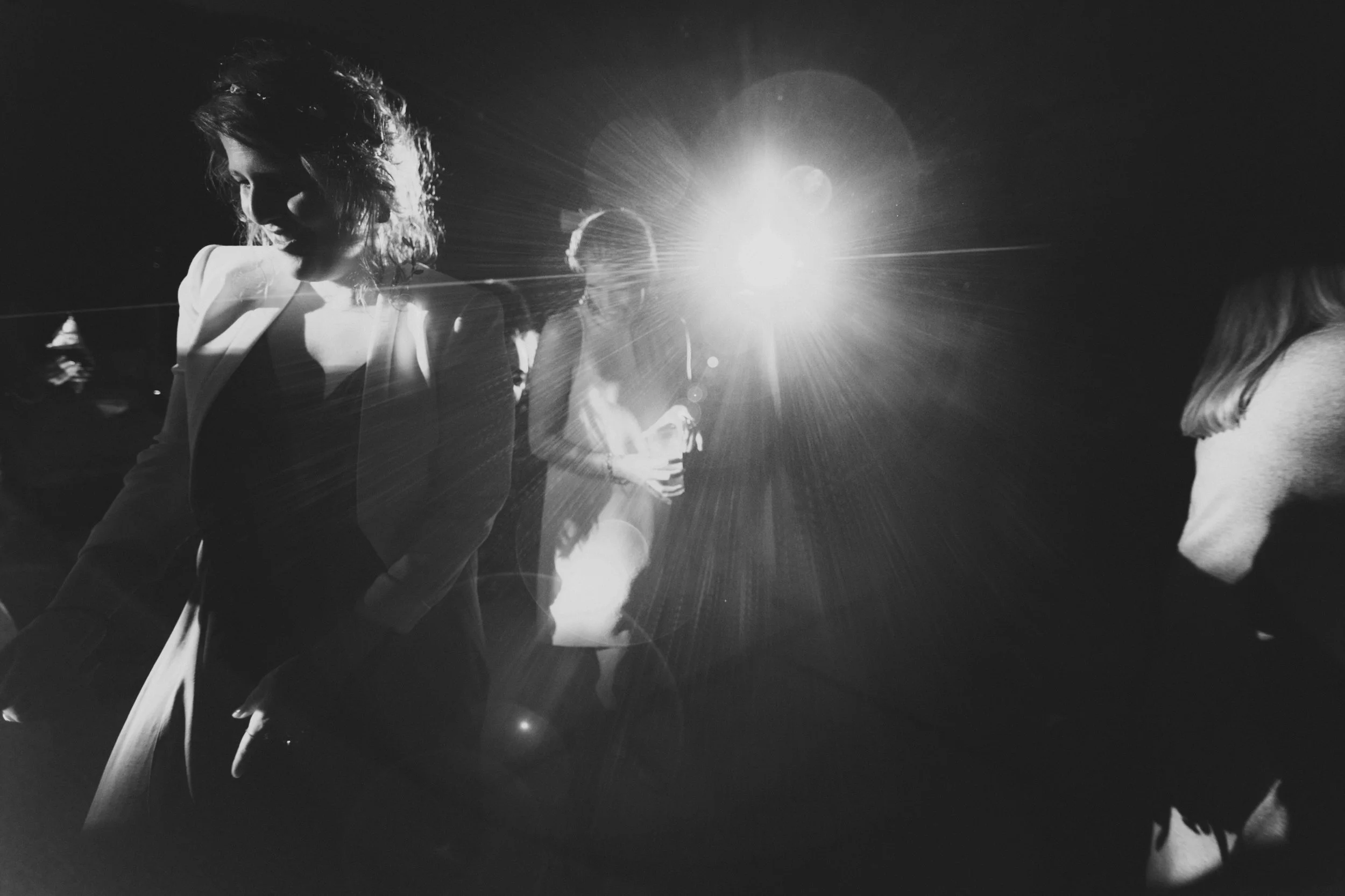 Black and white photo of people at a social event, with bright light source creating lens flare, some individuals engaged in conversation.