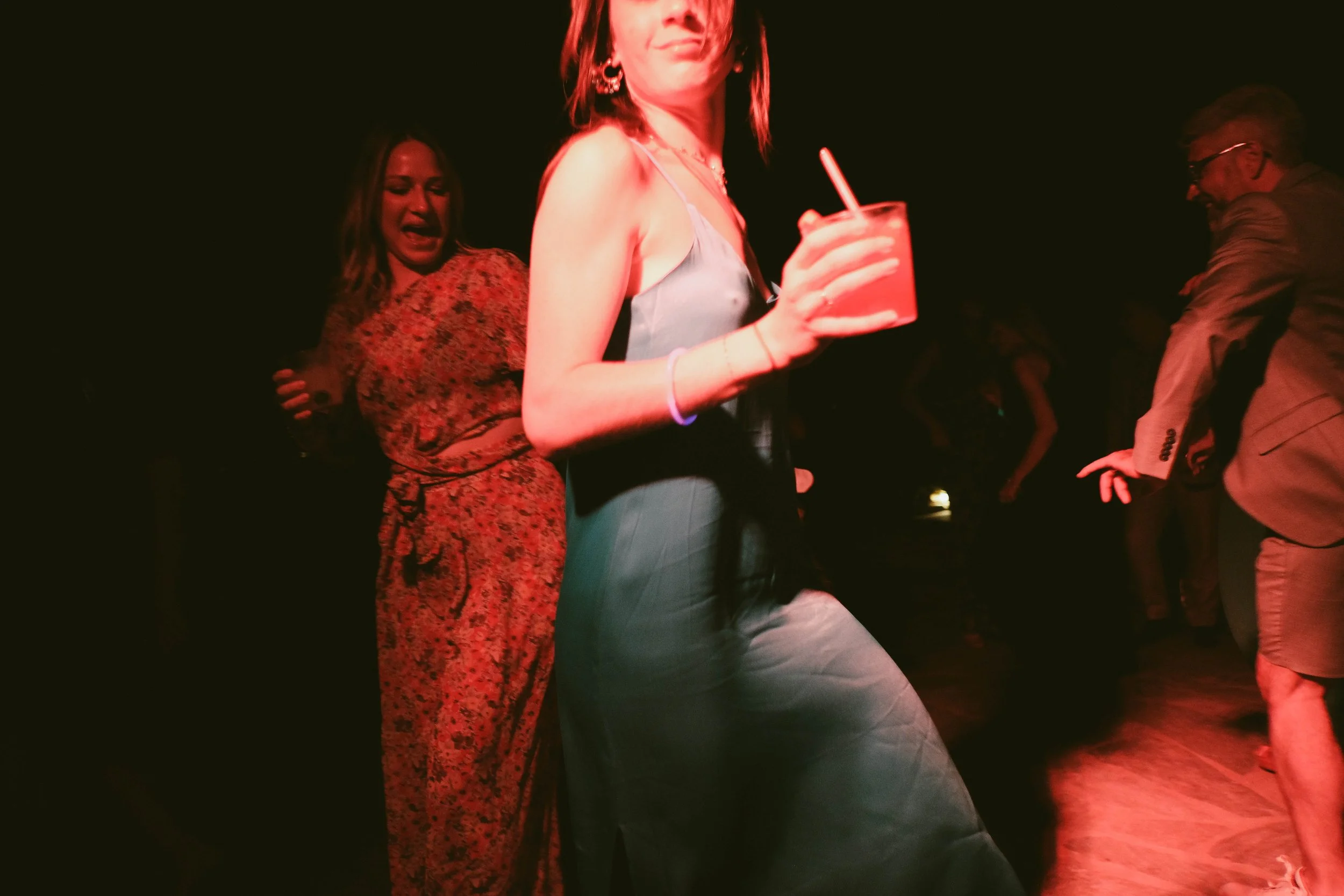 People dancing at a party, a woman in the foreground holding a drink, others in the background on a dance floor.