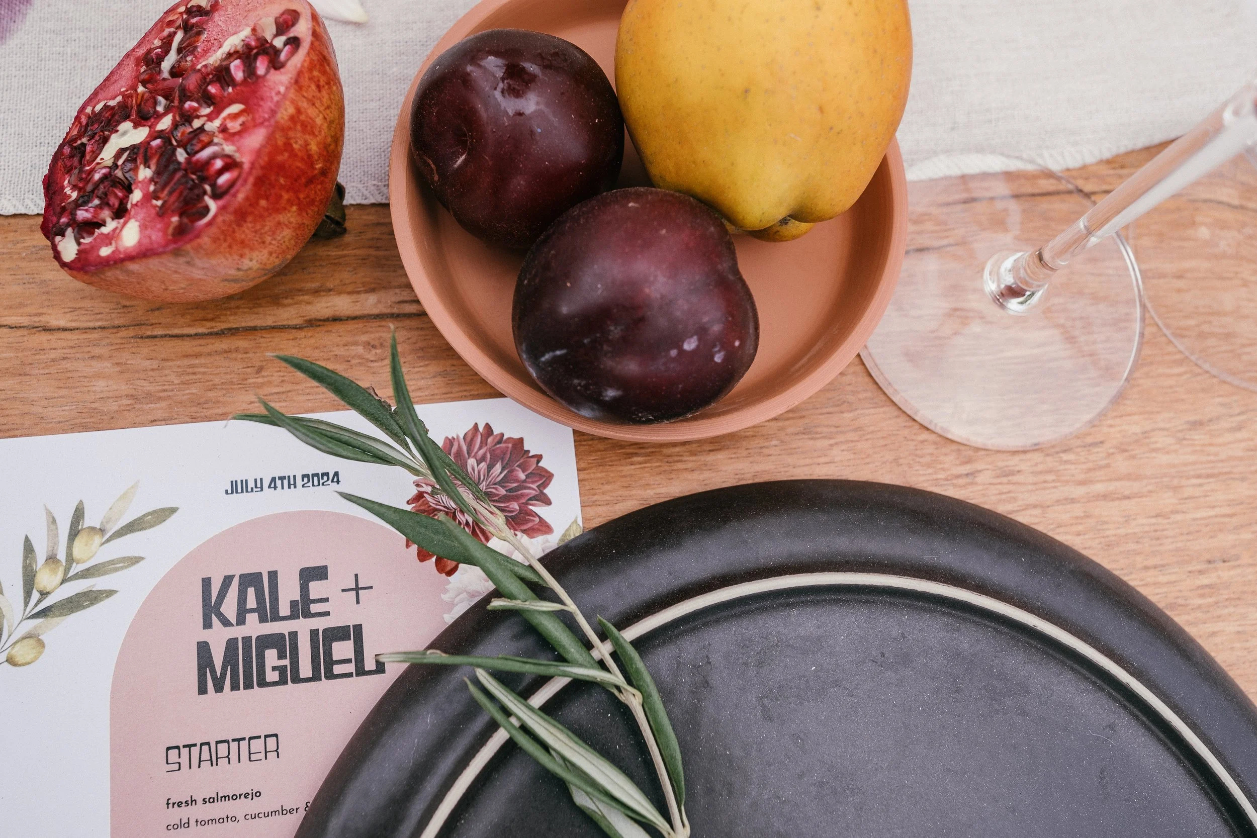 A table setting with a half pomegranate, a pink bowl with two plums and an apple, a wine glass with a white straw, a menu card for Kale + Miguel dated July 4th, 2024, and a black plate with a sprig of greenery on top.