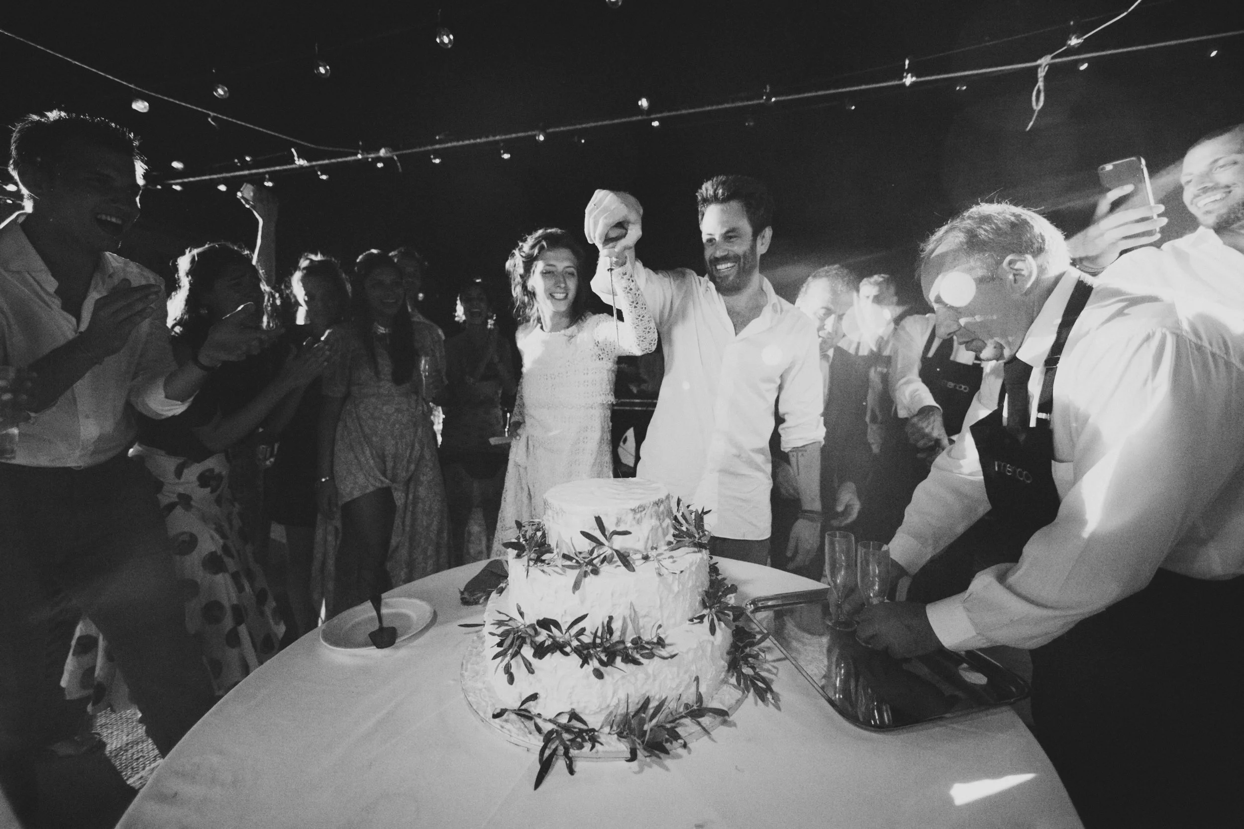 A group of people celebrating a wedding with a layered cake on the table, some clapping, and a man cutting the cake, under string lights at night.