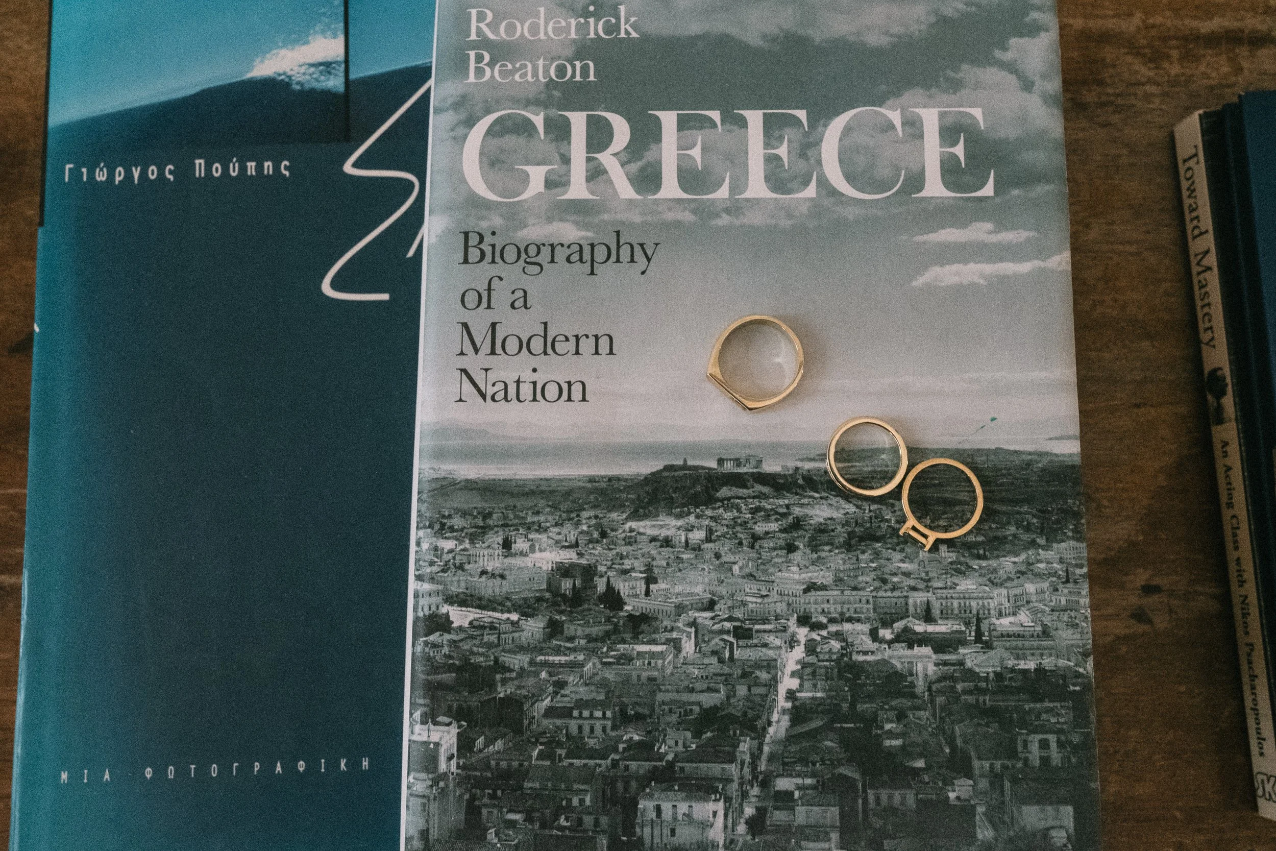 A black and white book cover featuring the cityscape of Greece with the title 'Greece: Biography of a Modern Nation' by Roderick Beaton, and three gold rings placed on top
