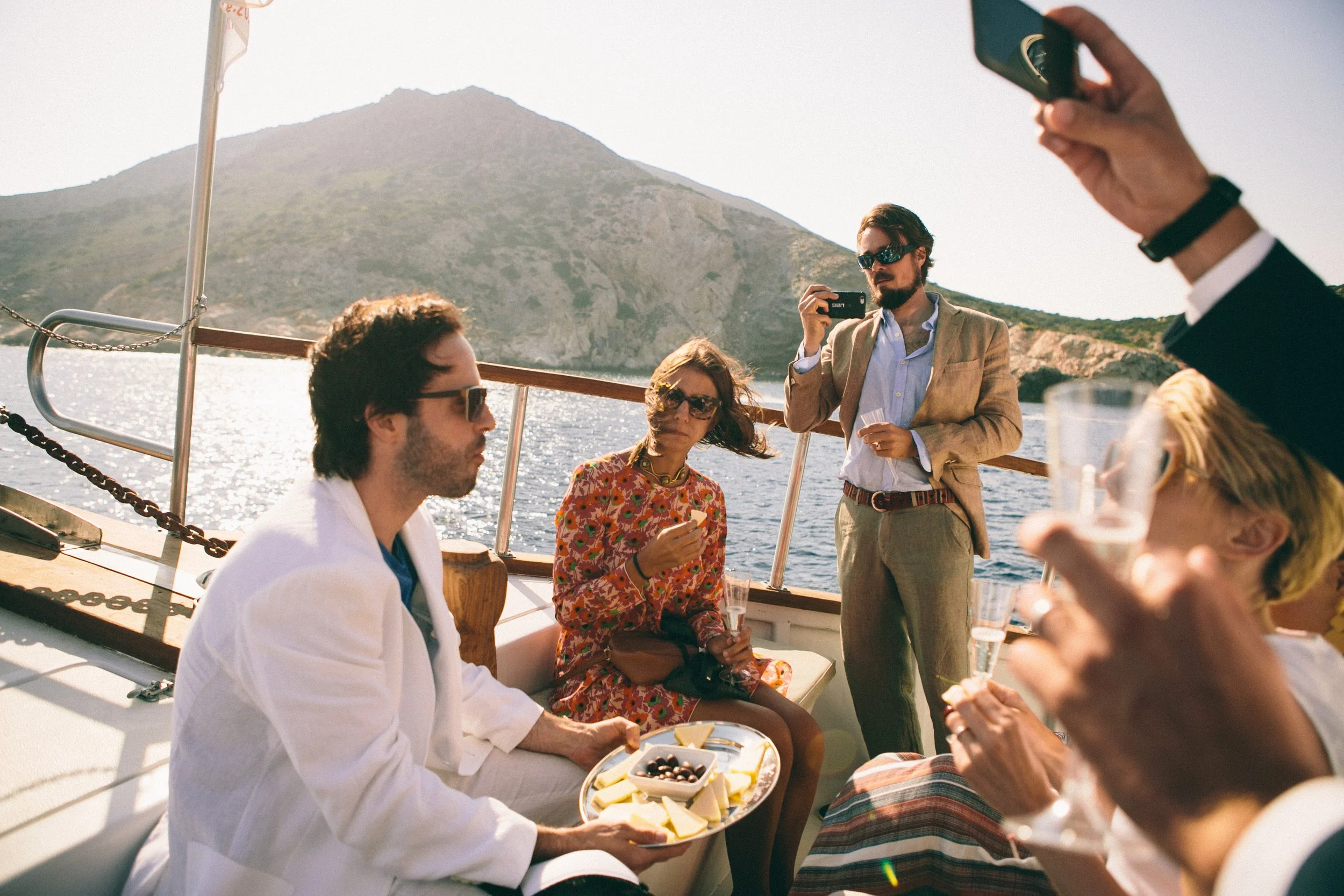 Group of people on a boat enjoying drinks and snacks, with a mountain and water in the background, some taking photos.