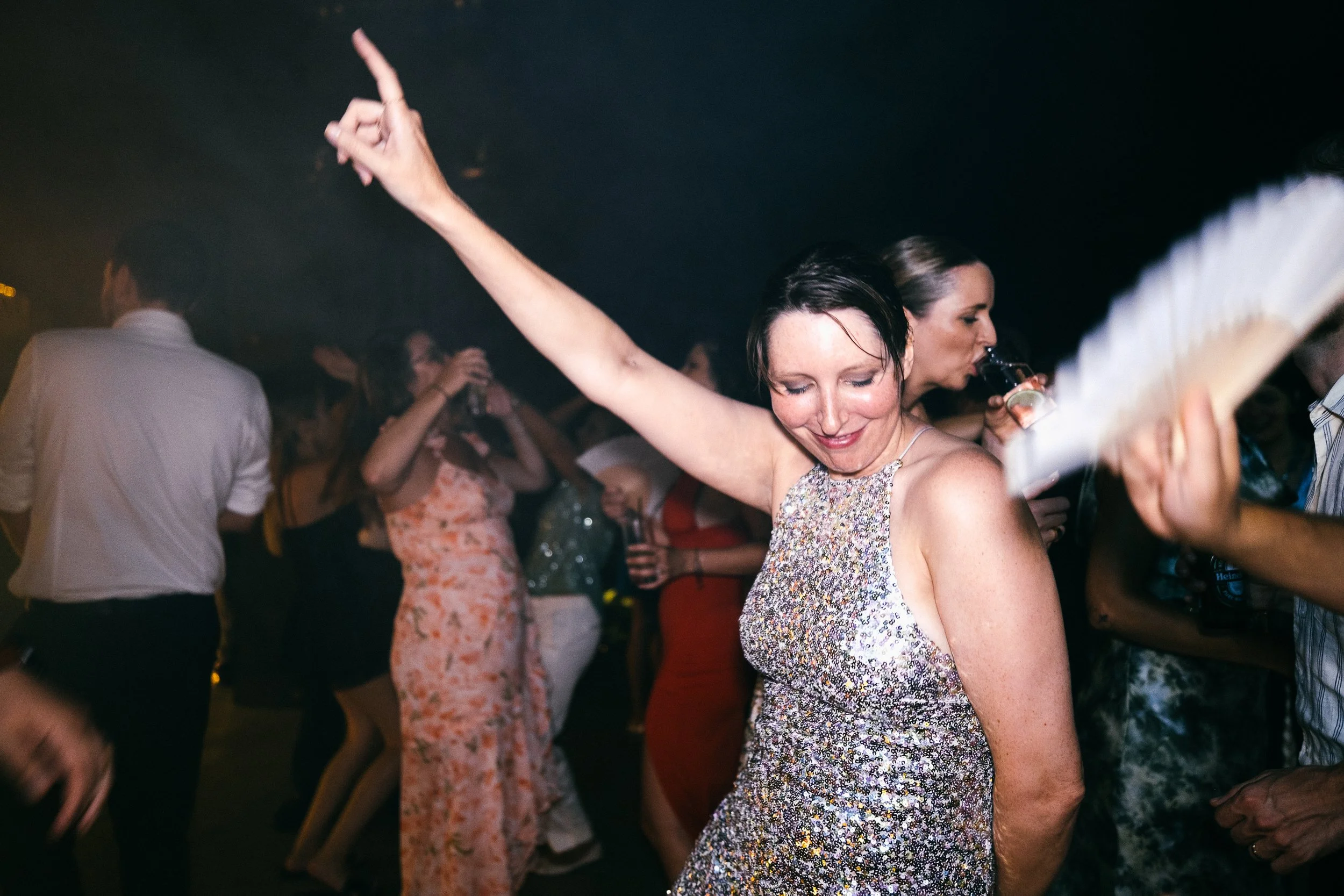 Woman dancing with her arm raised at a lively party with other guests in the background, some holding drinks.
