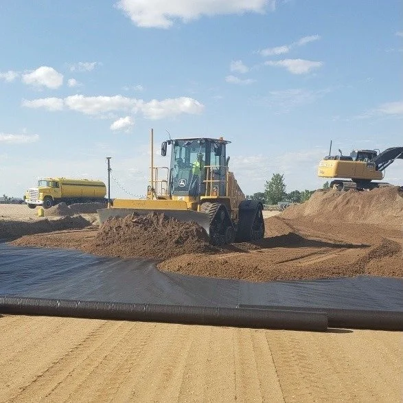 Construction site with a yellow road roller compacting soil, a water truck in the background, and an excavator on the right side of the dirt mound under a partly cloudy sky.