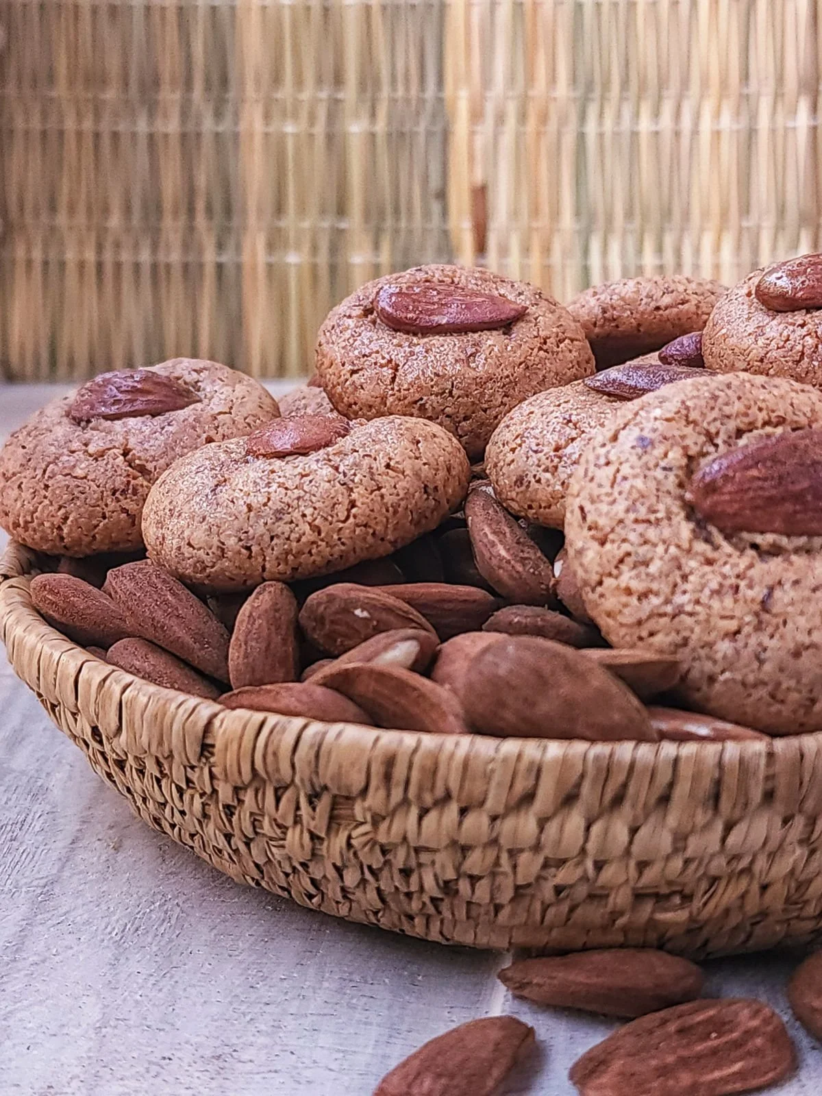 A woven basket filled with almond cookies topped with whole almonds, and some loose almonds around the basket, on a light-colored surface with a bamboo backdrop.
