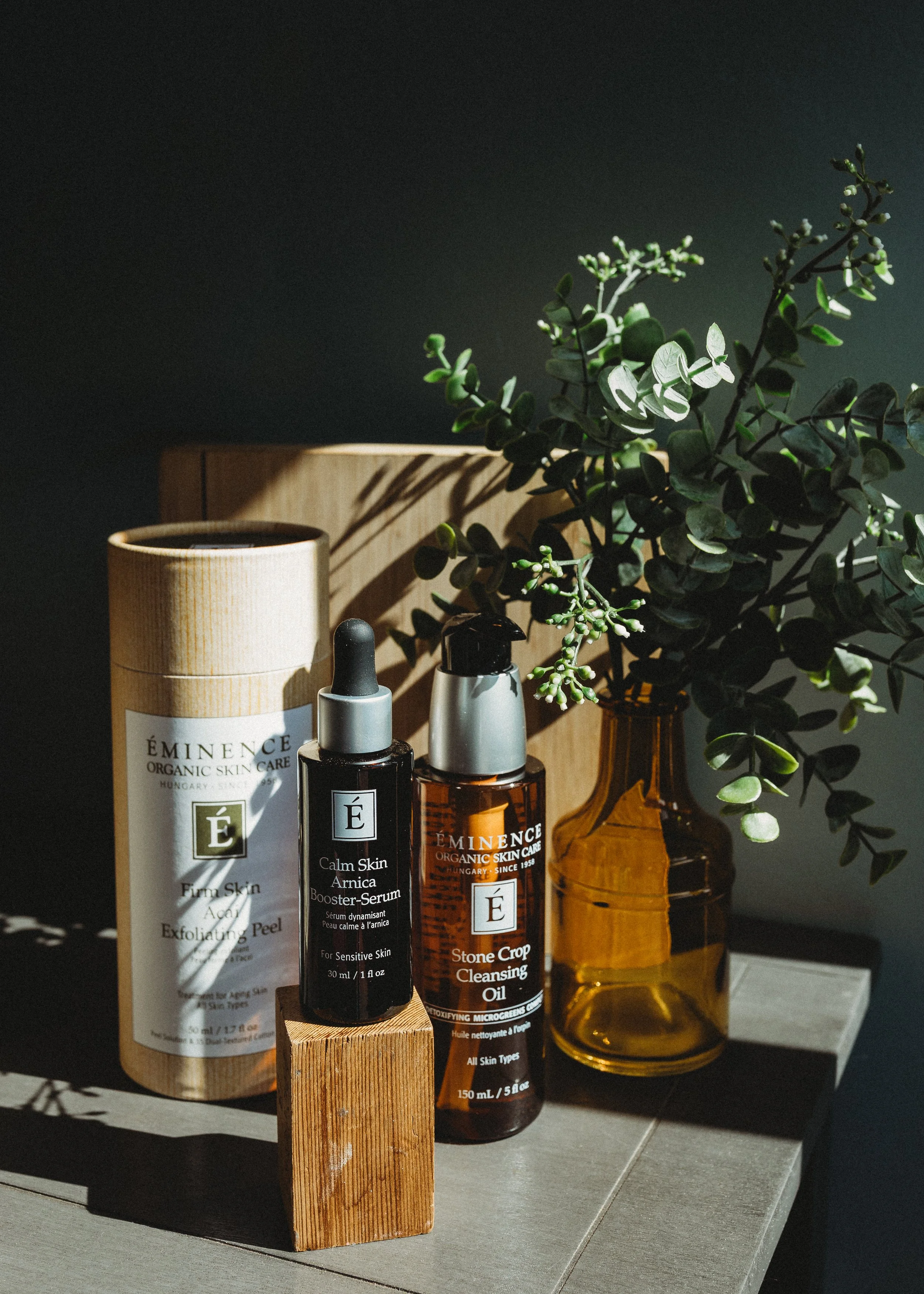 Éminence Organic Skin Care products including Stone Crop Hydrating Mist, Coconut Firming Body Lotion, Mangosteen Daily Resurfacing Cleanser, and Stone Crop Body Lotion arranged on a decorative surface with greenery.