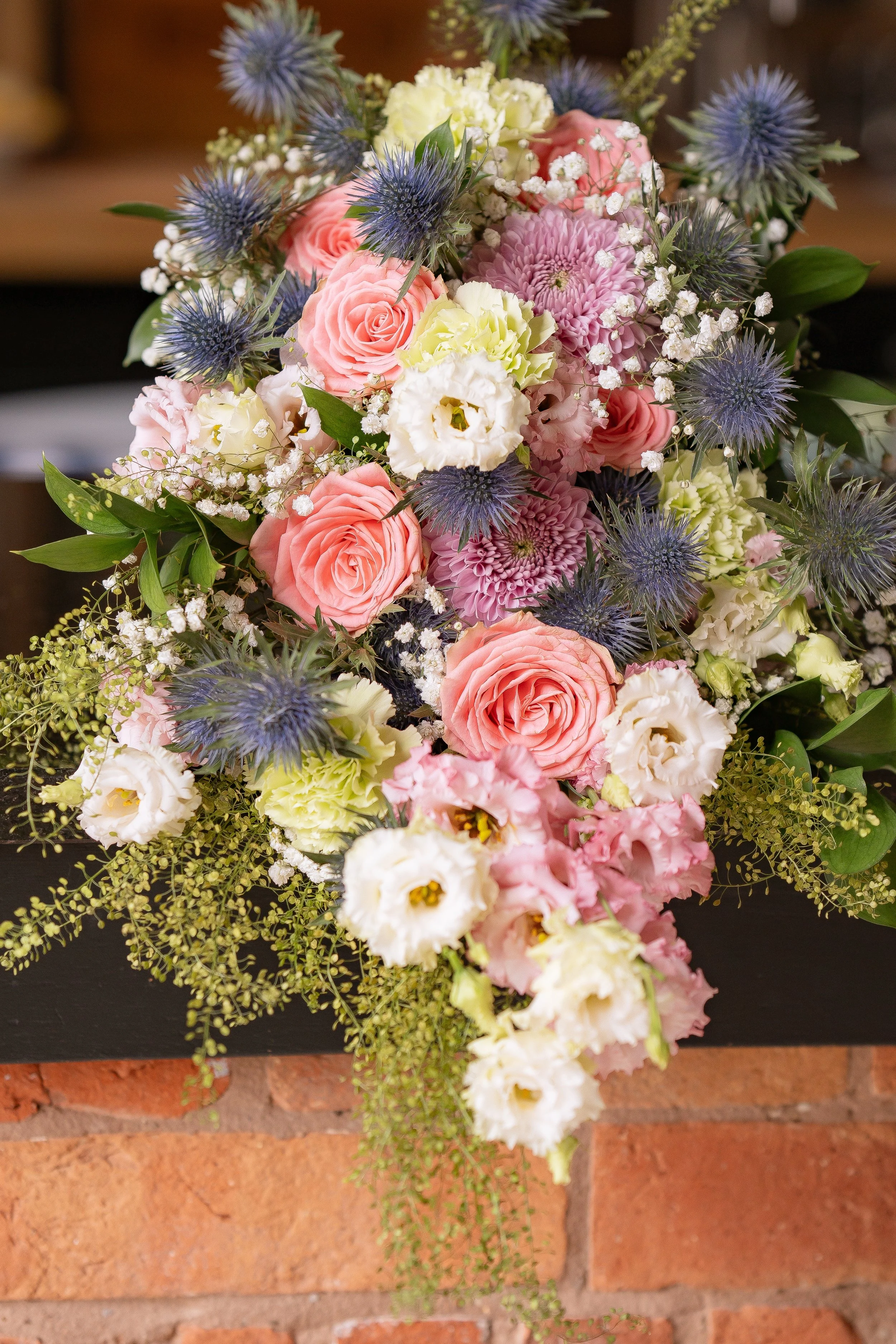 A cascading floral bouquet featuring pink roses, white lisianthus, purple thistles, lavender, and greenery.