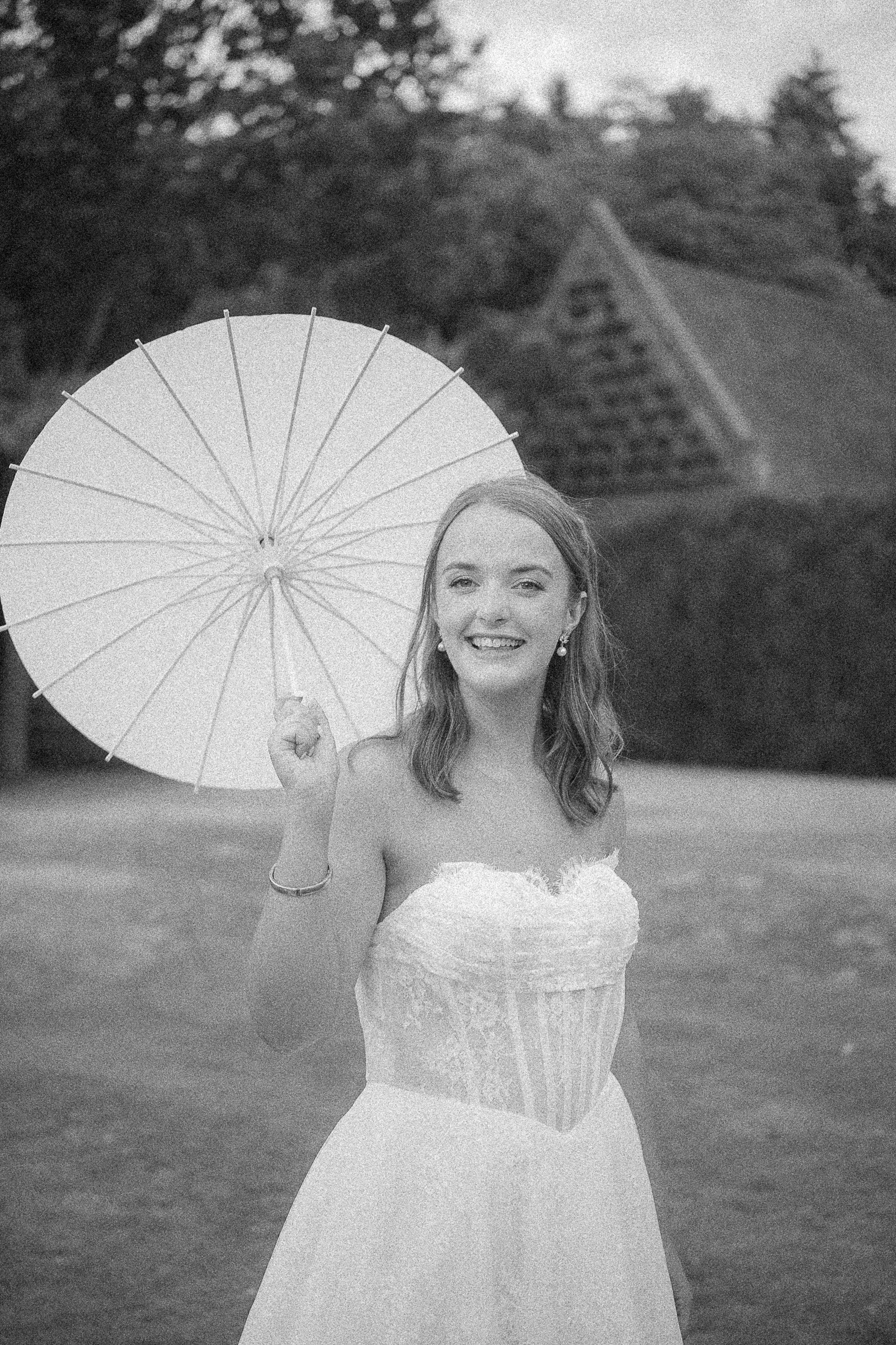 A young woman wearing a strapless wedding dress, holding a white parasol, smiling outdoors in a grassy area with trees in the background.