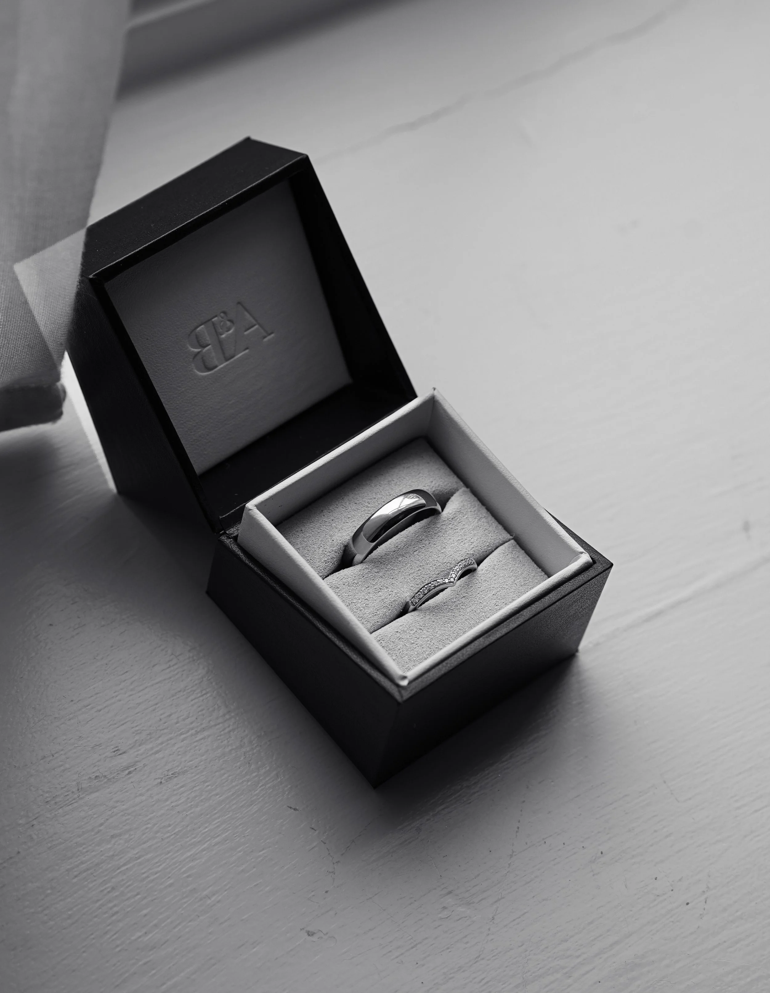 A black jewelry box with a white interior, containing a silver ring and a silver band with small gemstones, placed on a white surface.